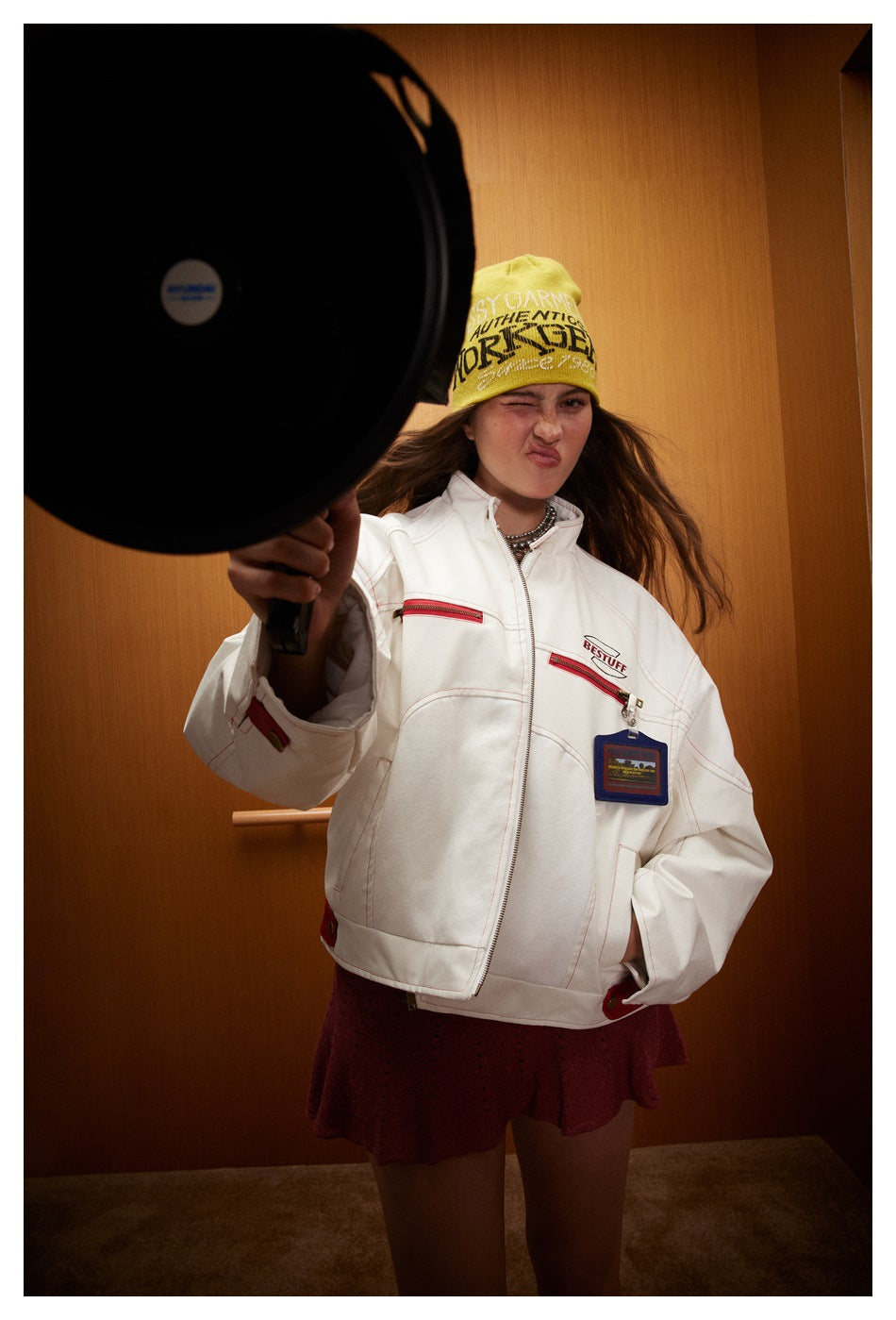 Model wears a white jacket with red zip pockets and a burgundy skirt; a large black prop partly obscures the jacket.