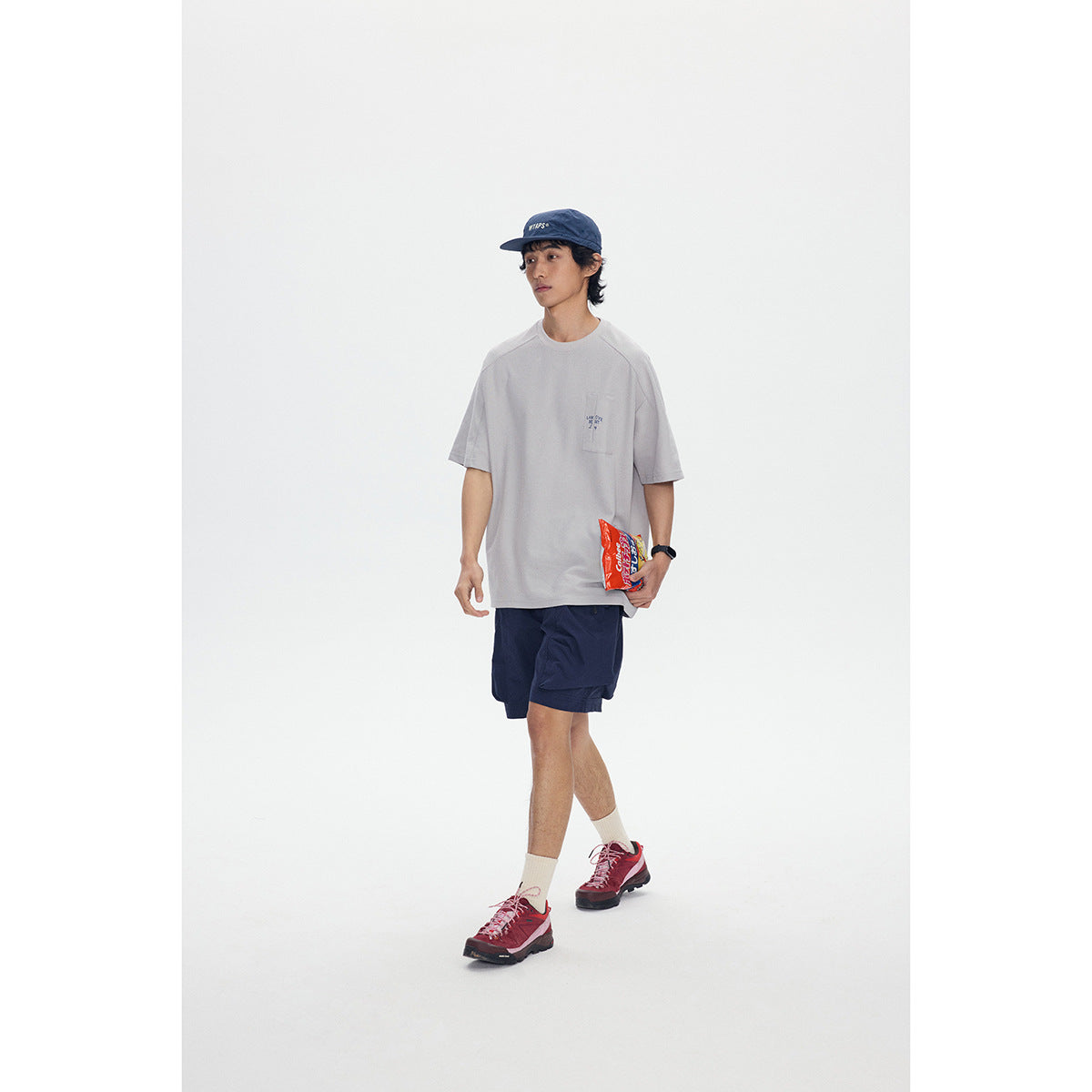 Full-length model wearing a light gray pocket T-shirt, navy shorts, and a navy cap.