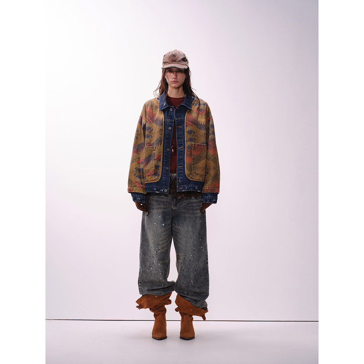 Model wearing a patterned brown, rust and navy jacket with denim trim, baggy jeans and brown boots, front view.