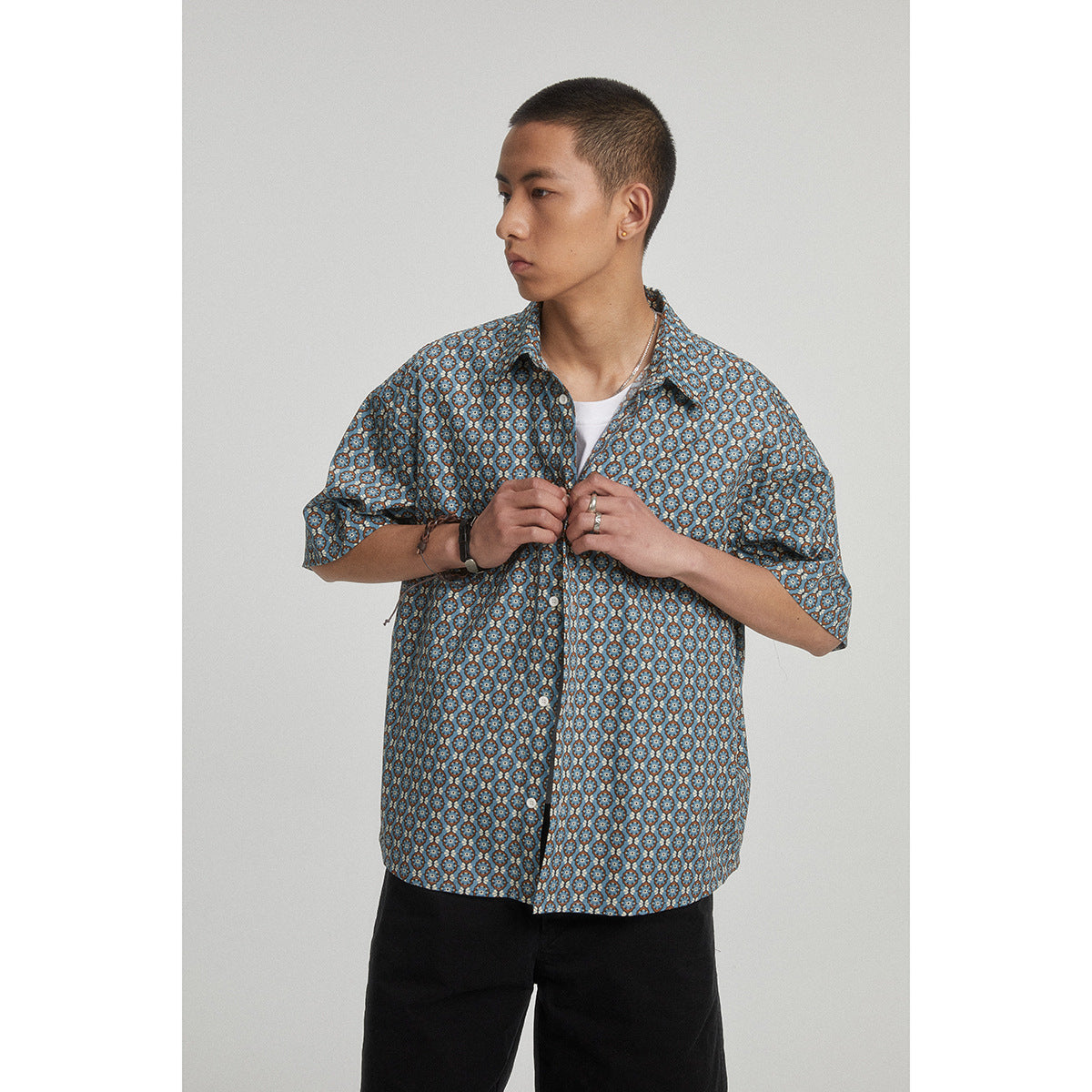 Model wearing a blue geometric-pattern short-sleeve button-up over a white tee, shown fastening the shirt.