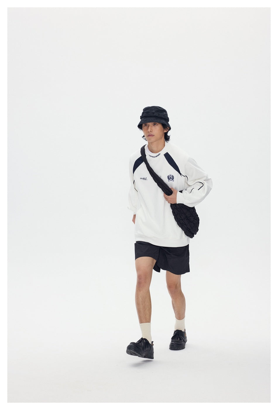 Full-length model wearing a white sweatshirt with navy accents, black shorts, and a black shoulder bag.