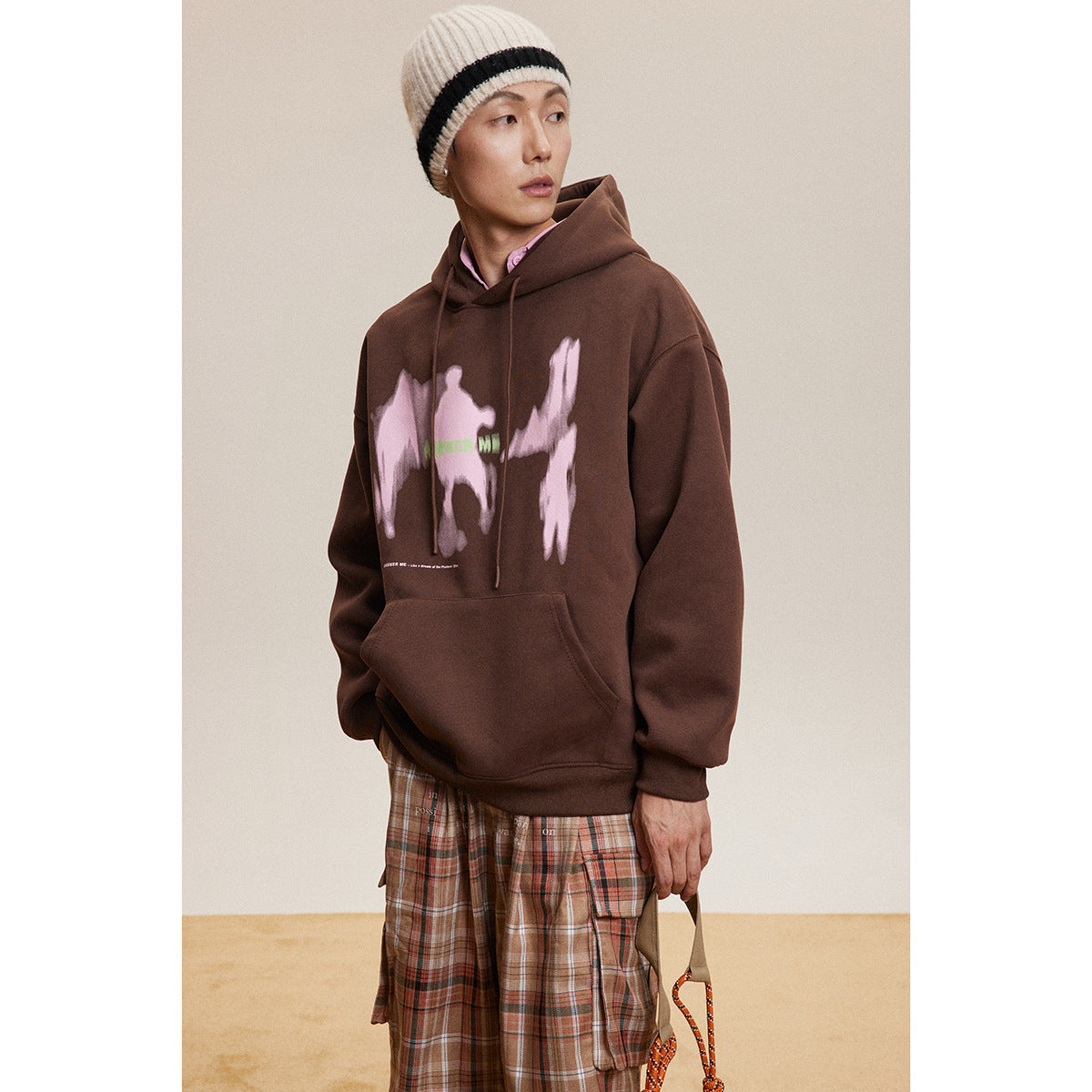 Standing model in a dark brown hoodie with a pink abstract chest graphic and plaid cargo trousers.