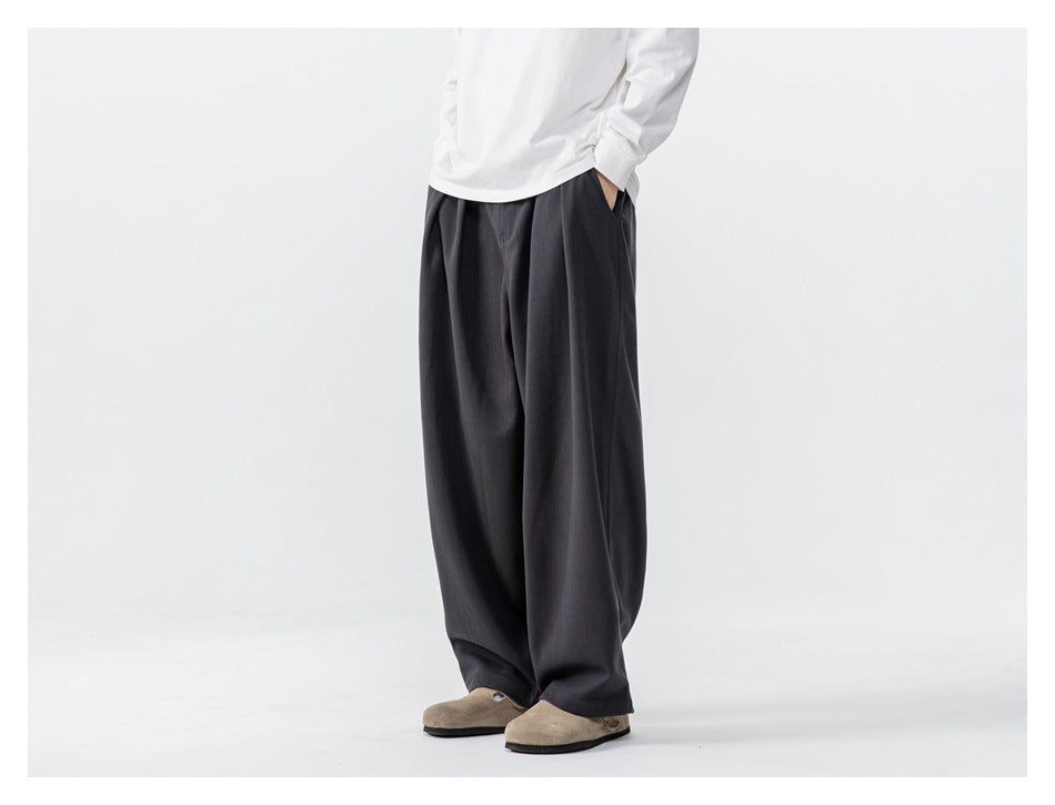Person wearing dark gray wide-leg pleated trousers with a white top and tan clogs, three-quarter view, hand in pocket
