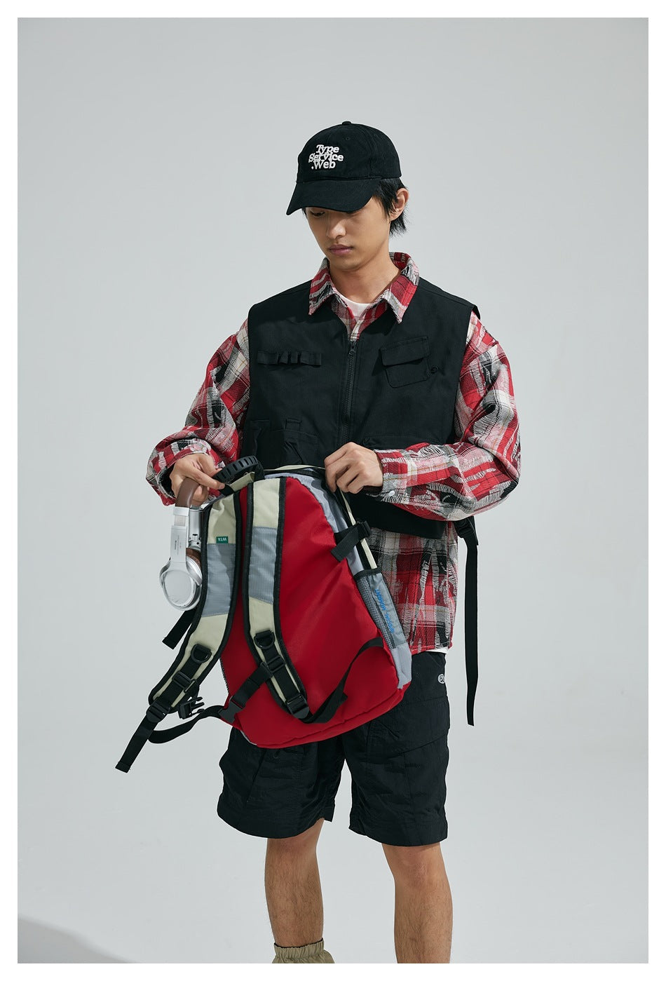 Model wearing a black zip-front utility vest over a red plaid shirt, holding a backpack and headphones