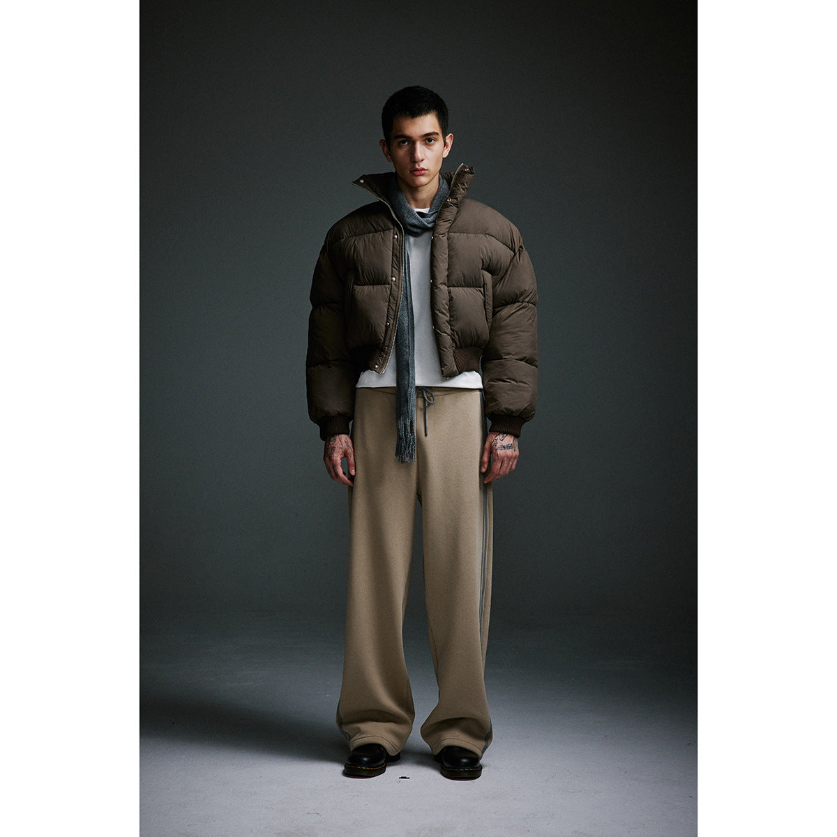 Model wearing a dark brown puffer jacket, gray scarf, white shirt and loose taupe trousers.