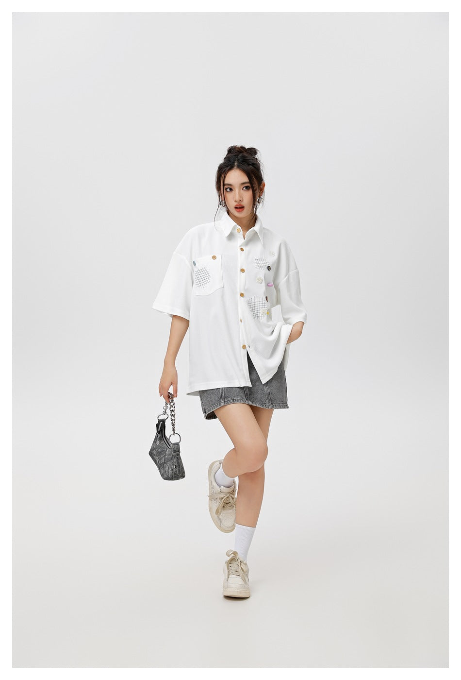 Model wearing a white oversized short-sleeve button-up shirt with grid patches and colorful accents, paired with gray shorts.