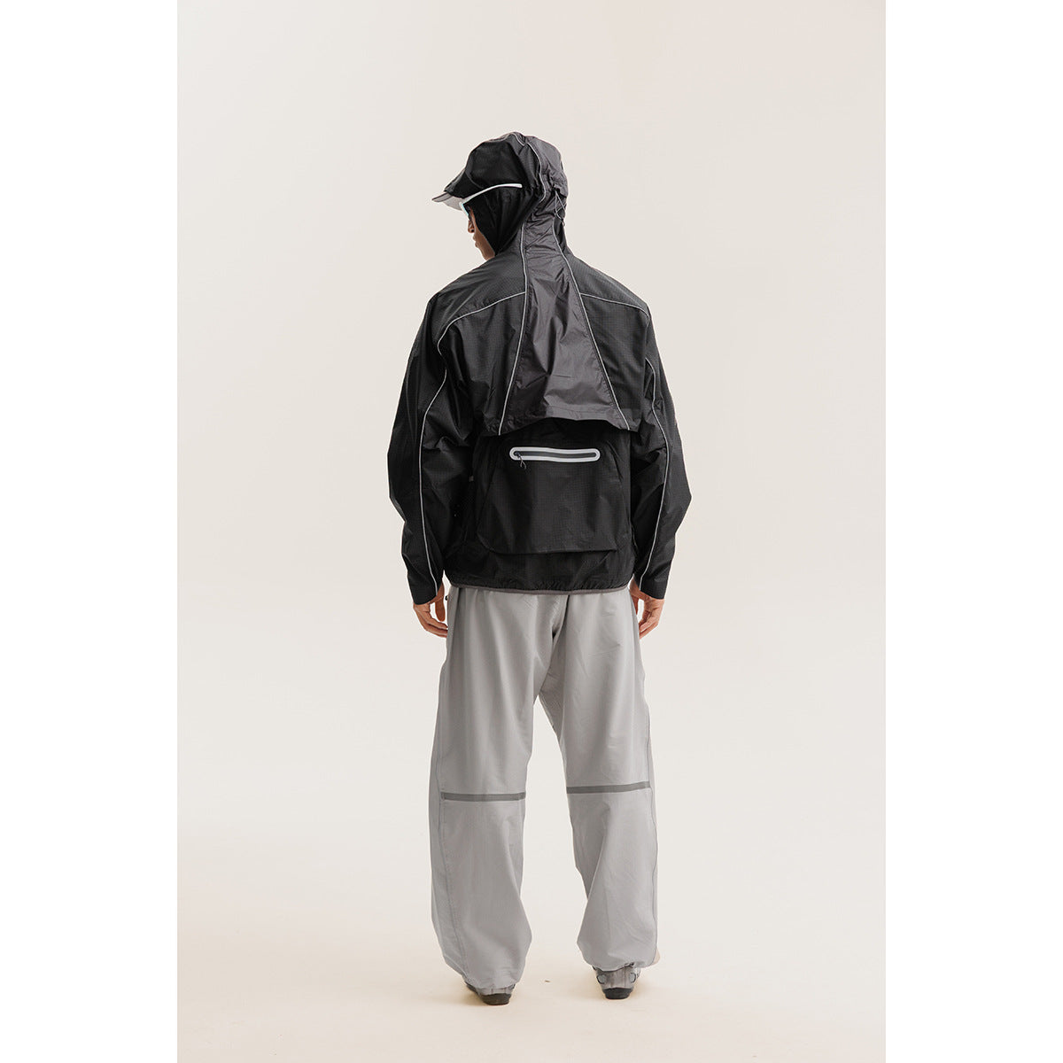 Rear view of a model wearing a black hooded jacket with reflective piping and a back pocket, plus gray pants.