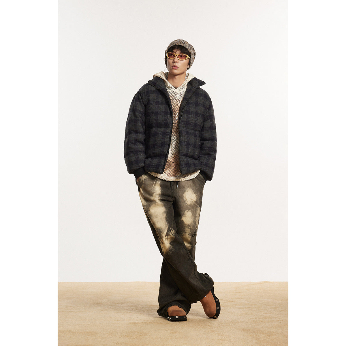 Model wearing a dark plaid puffer jacket over a cream open-knit hoodie and mottled brown wide-leg pants.