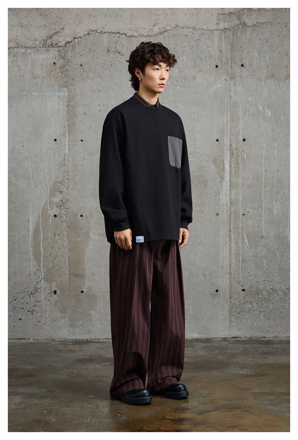 Model wearing a black oversized crewneck sweatshirt with a gray chest pocket and wide striped trousers.