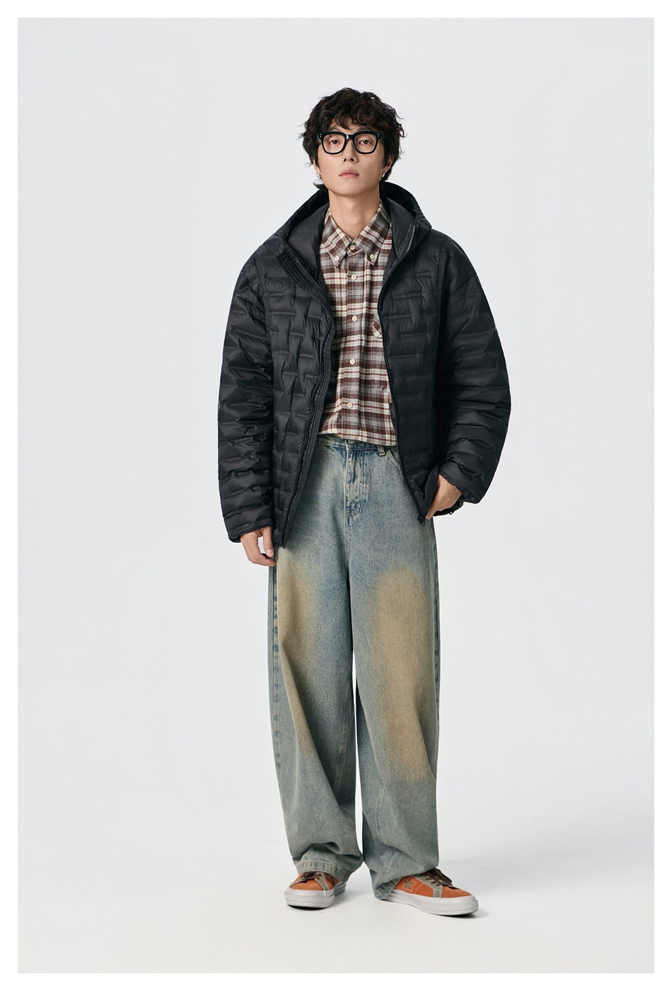 Model wearing an open black quilted hooded puffer jacket with a plaid shirt and loose faded jeans.