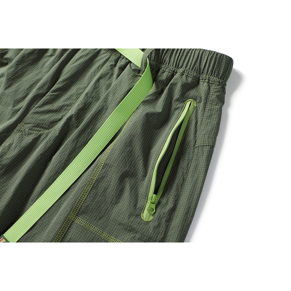 Close-up of olive grid-pattern trousers showing an elastic waistband, lime belt and vertical zip pocket.