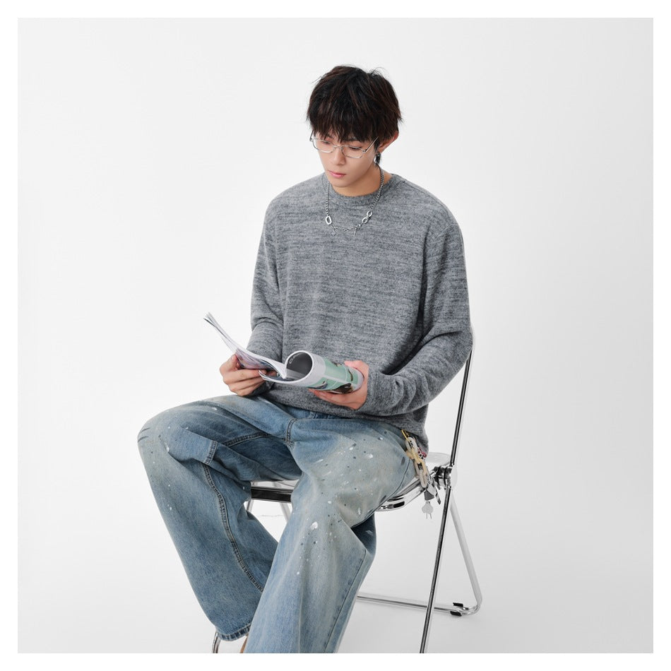 Seated model wearing a marled gray crew-neck sweater and light-wash jeans while reading a magazine.