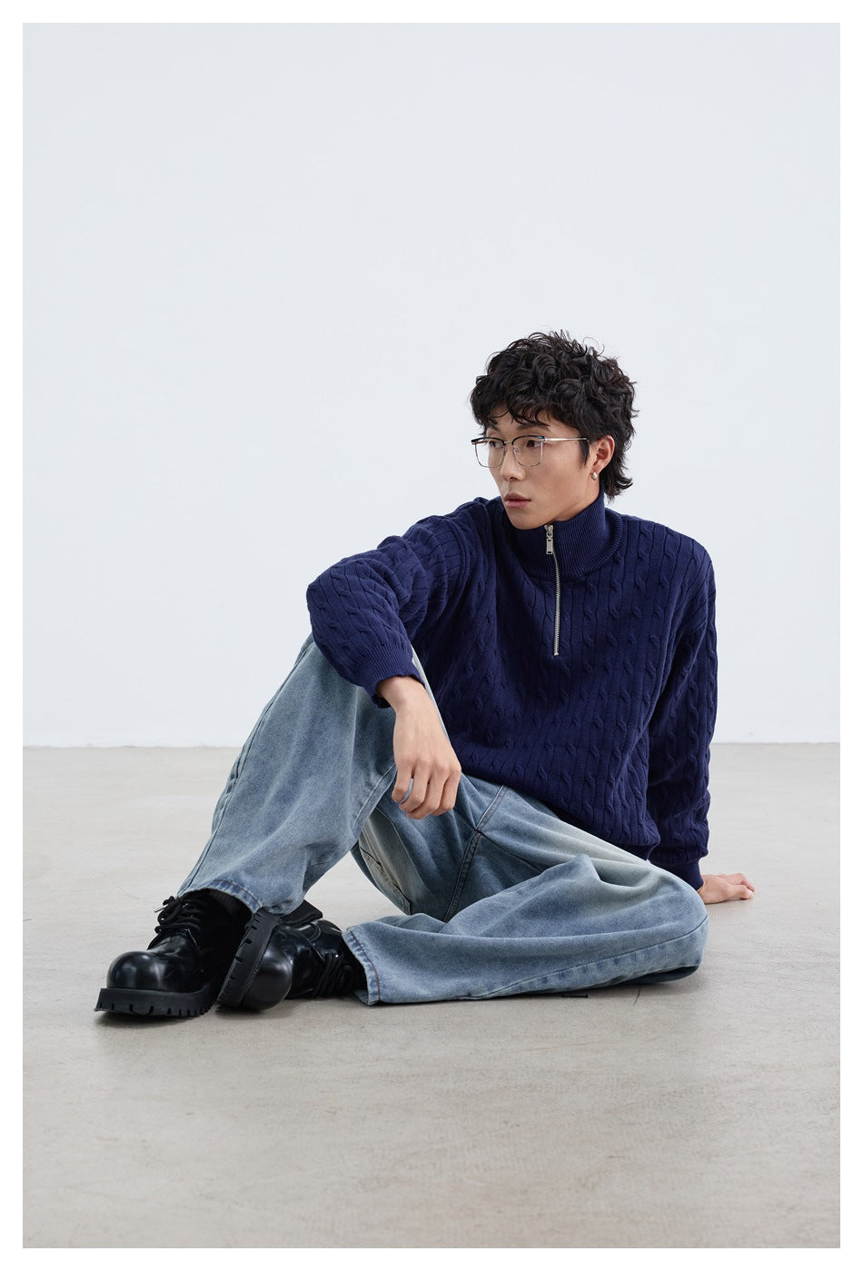 Model seated wearing a navy cable-knit sweater with a high zip collar and light blue jeans.