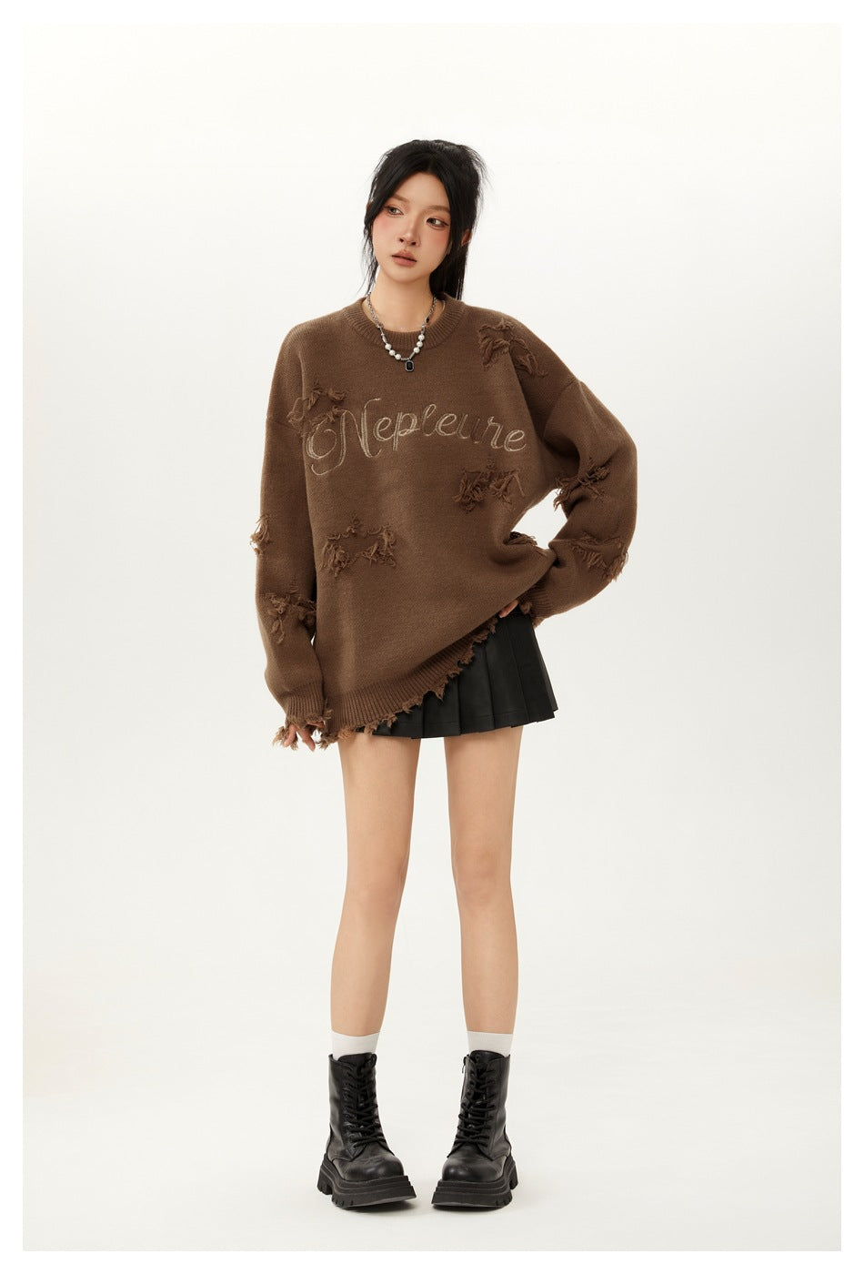 Model wearing an oversized brown distressed sweater with decorative chest lettering, a black pleated skirt and lace-up boots.