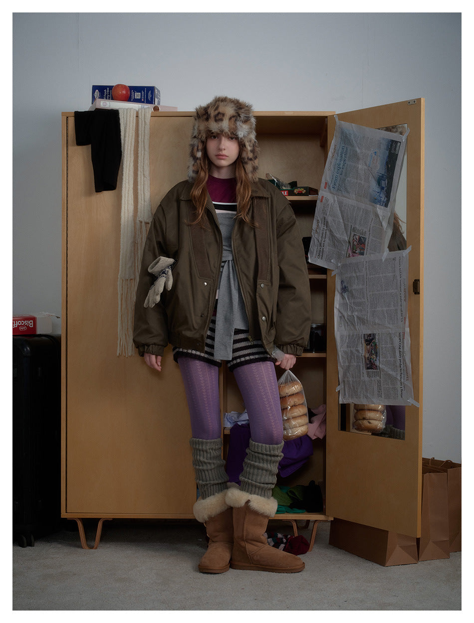 Model wearing the olive padded-look jacket open over a layered outfit, standing in front of a wooden wardrobe