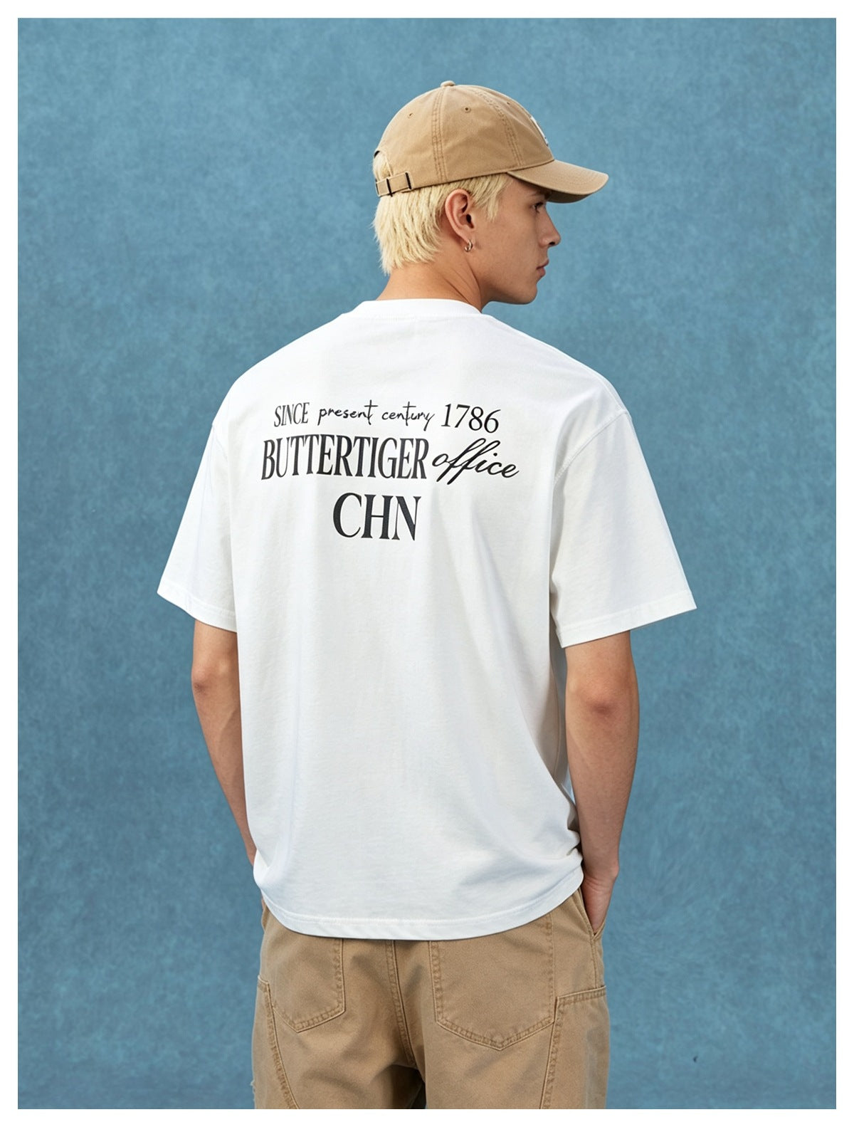 Person seen from behind wearing a white T-shirt with black lettering print, tan cap and khaki pants against a blue backdrop