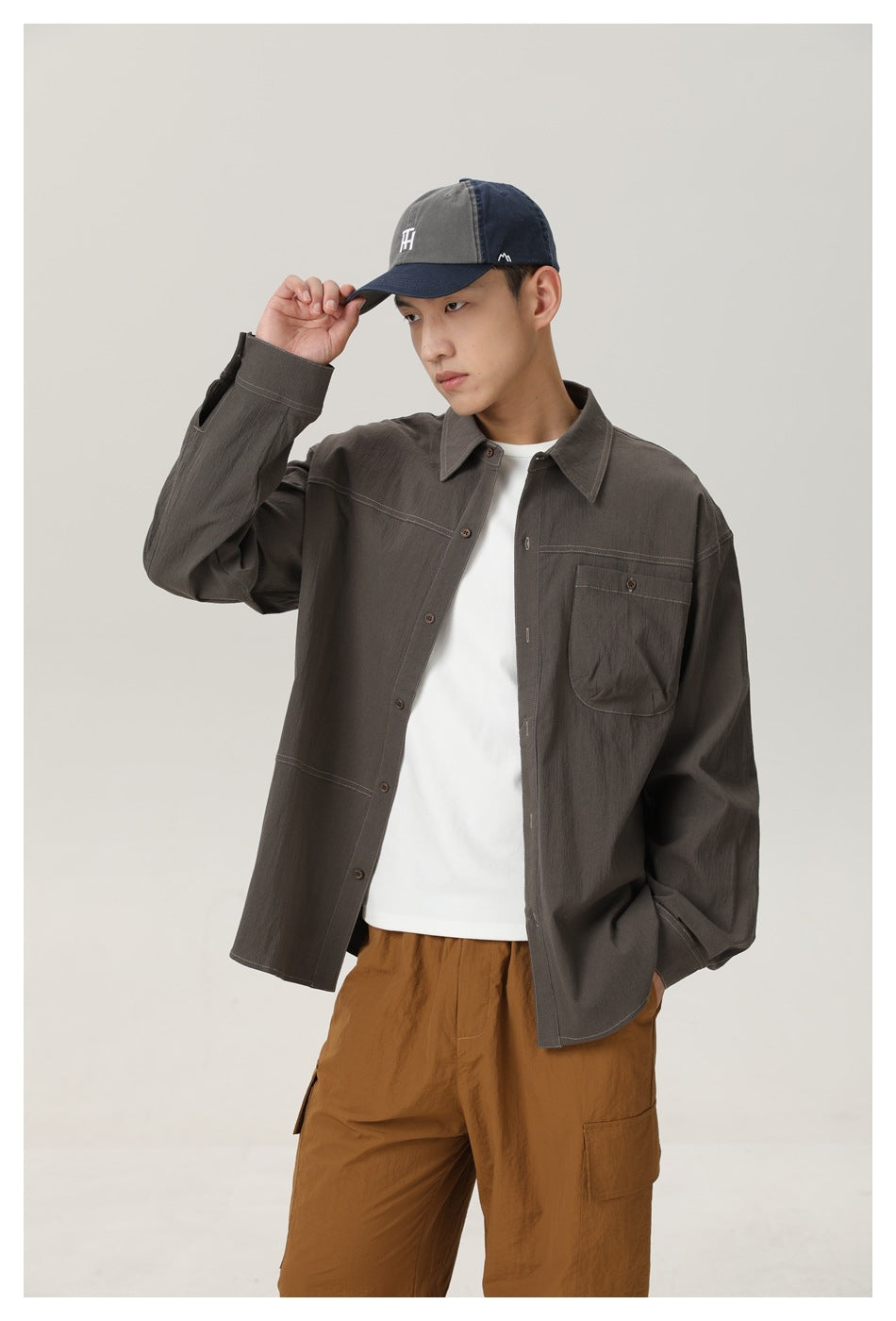 Man in gray-green button-up shirt over white tee with chest pocket and contrast stitching, hand on cap brim