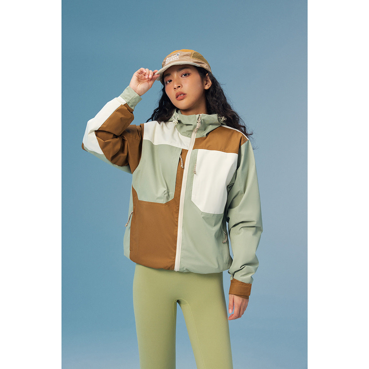 Model wearing a sage, cream, and brown color-block hooded jacket with green leggings and a tan cap.