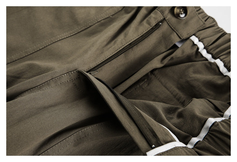 Close-up of olive-brown trousers showing a zippered opening, buttoned waistband tab, and white trim.