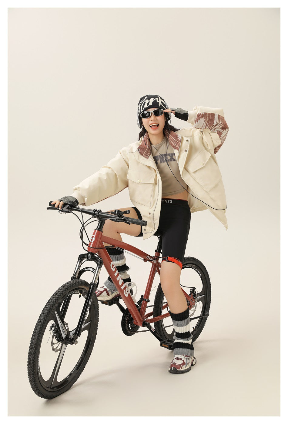 Model wearing an open cream jacket with rust patterned collar and sleeve panels while posing on a bicycle.
