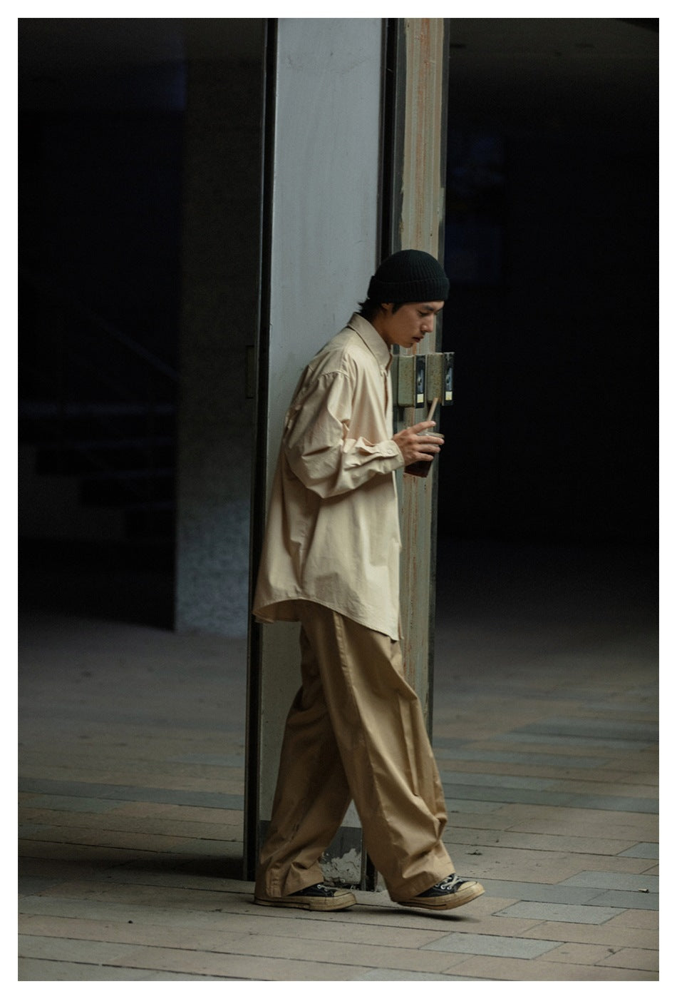 Model wearing an oversized light beige button-up shirt, loose tan trousers and a black knit beanie.