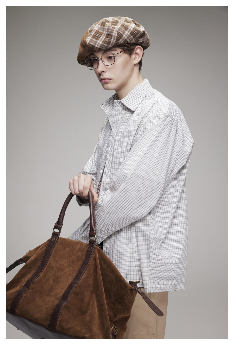 Close-up of a model wearing an oversized light gray checked button-up shirt and brown plaid cap, holding a brown bag.