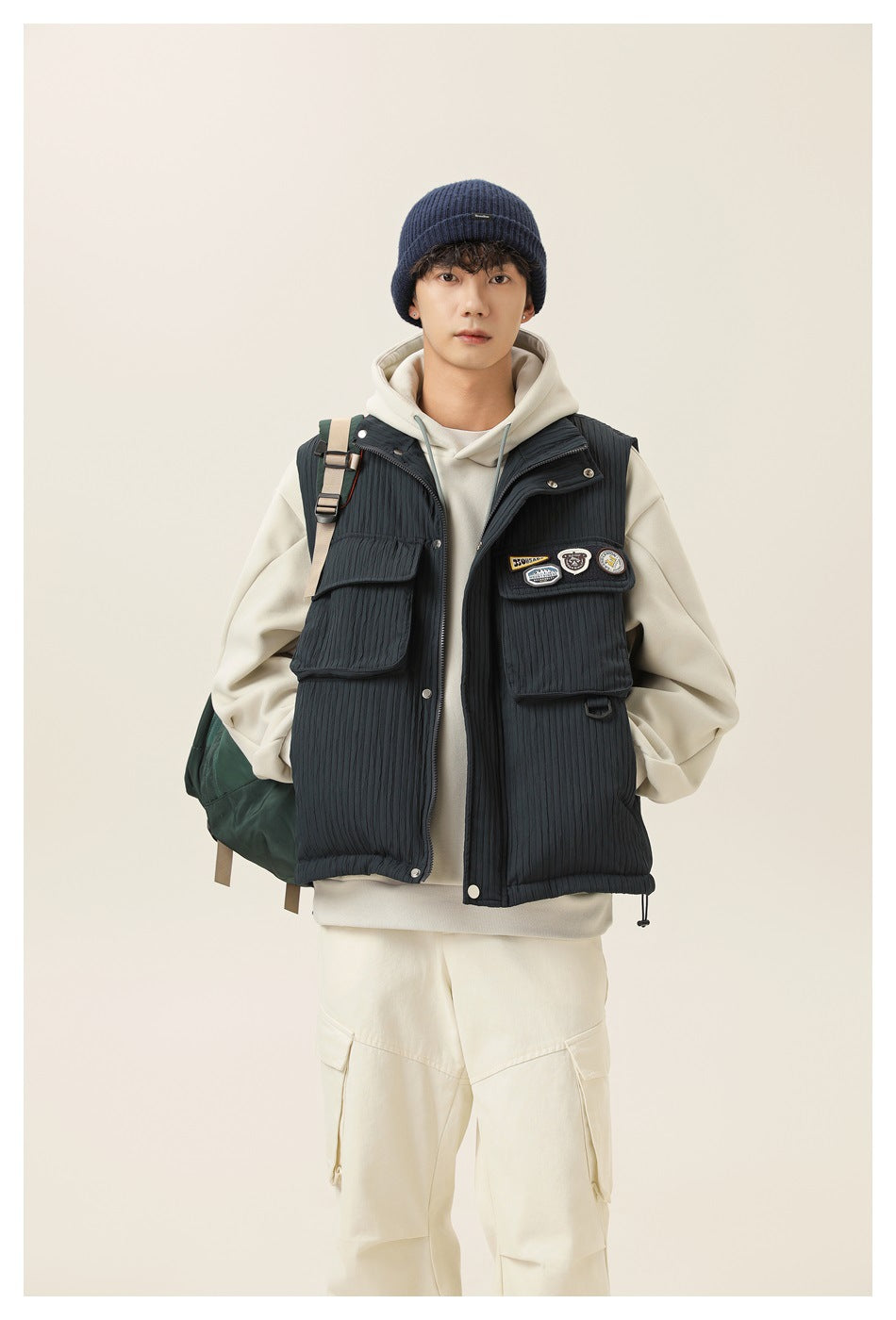 Front view of a model wearing a dark charcoal quilted utility vest with large flap pockets over an ivory hoodie.