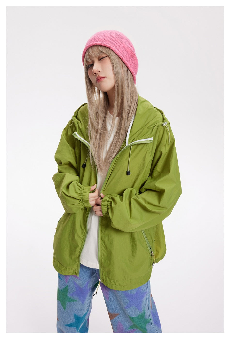 Model wearing an open lime-green hooded jacket over a white shirt, with a pink beanie and patterned jeans.