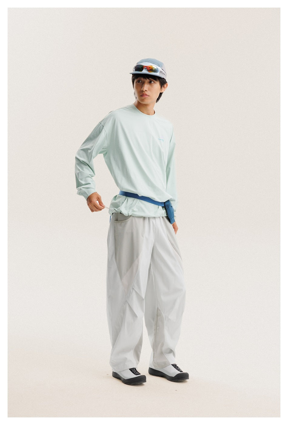 Model wearing a pale mint long-sleeve shirt and loose white trousers, with a blue waist strap.