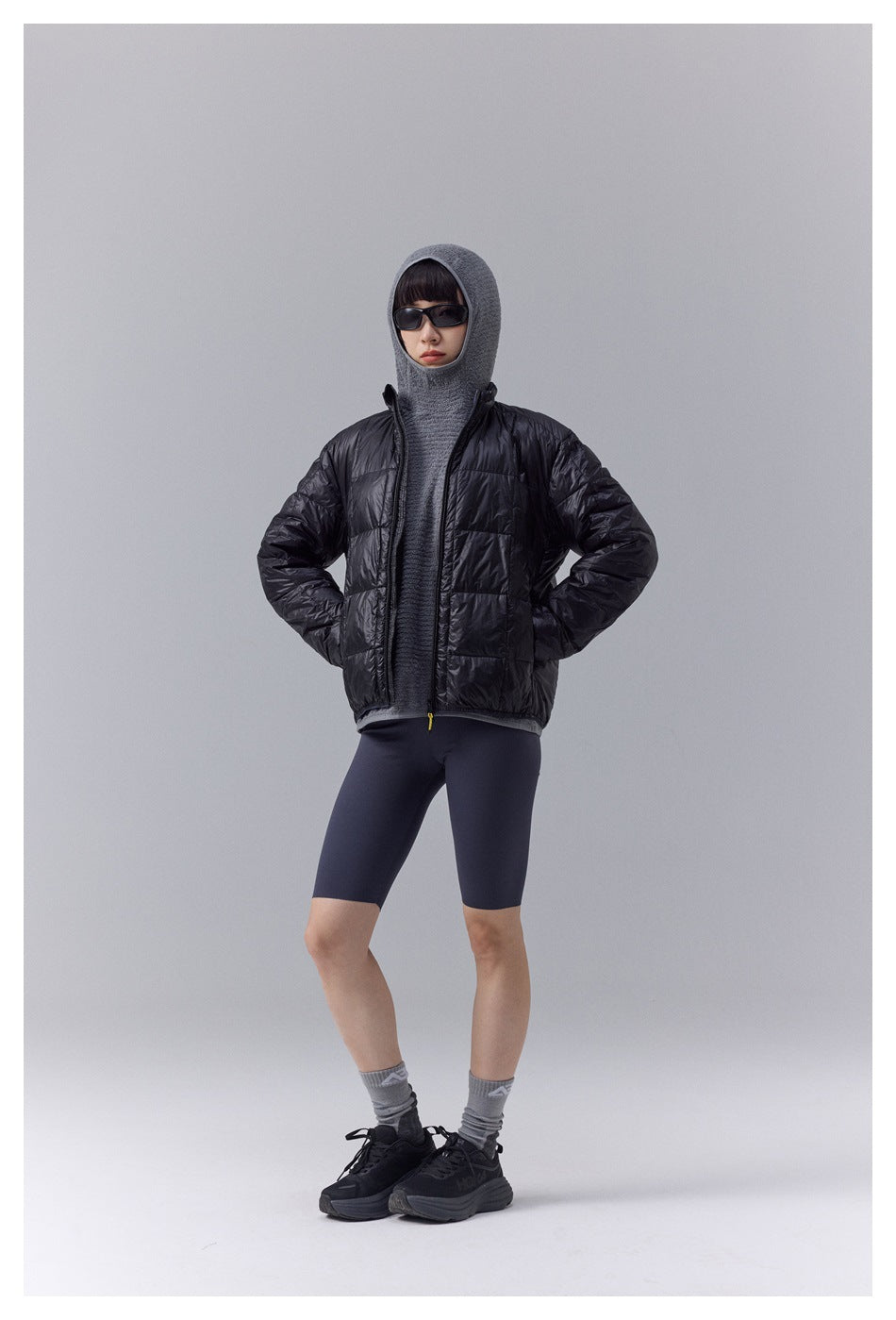 Model wearing black quilted puffer jacket over gray knit hood and top, with dark shorts, front view