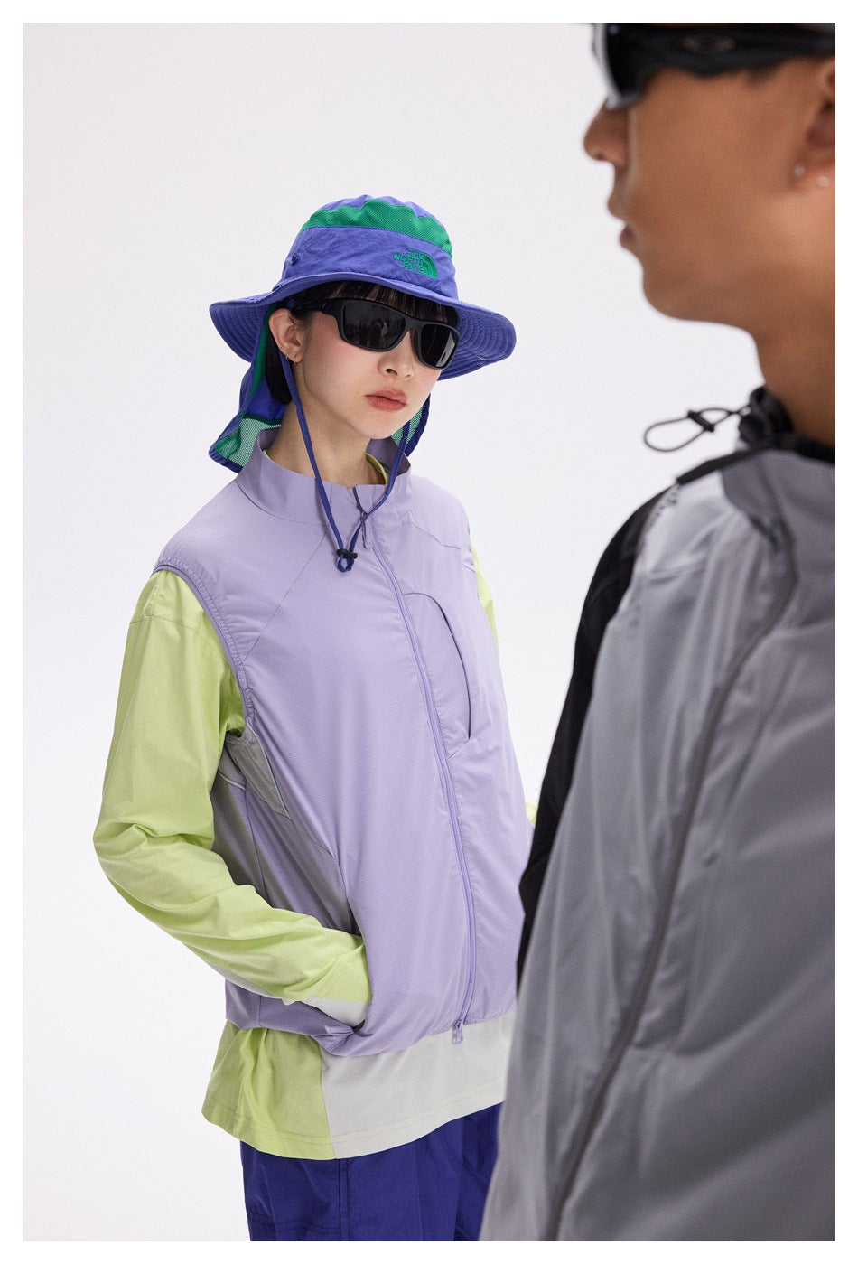 A model wears a lavender zip vest over a lime long-sleeve top and blue shorts; another model is partly visible in front.