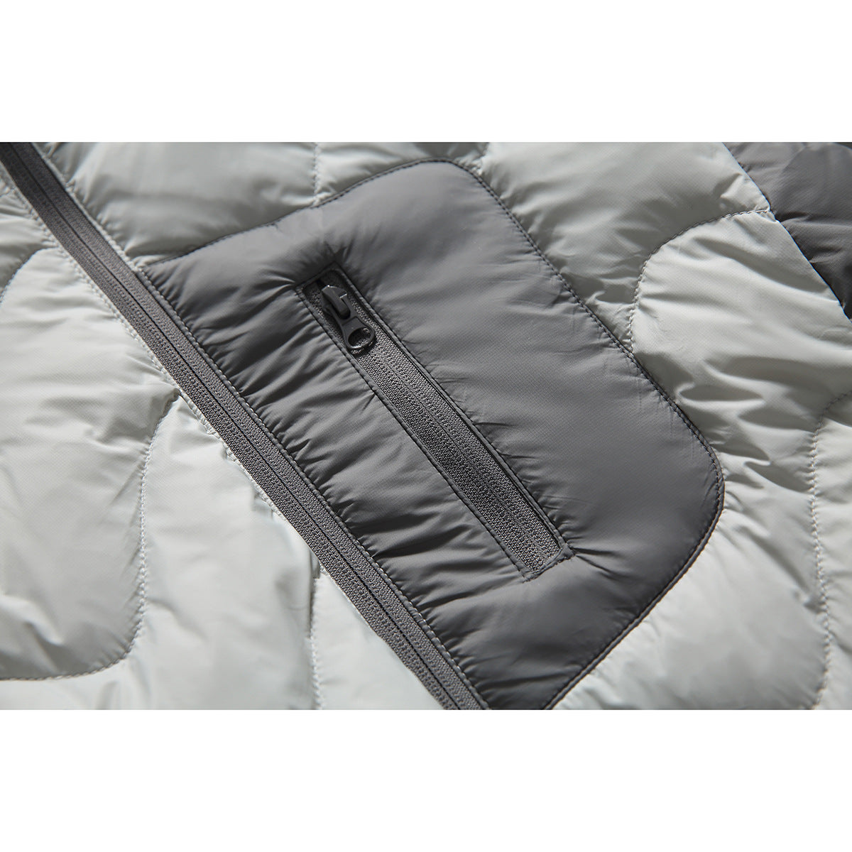 Close-up of the dark gray zippered chest pocket and front zipper on a light gray quilted puffer jacket.