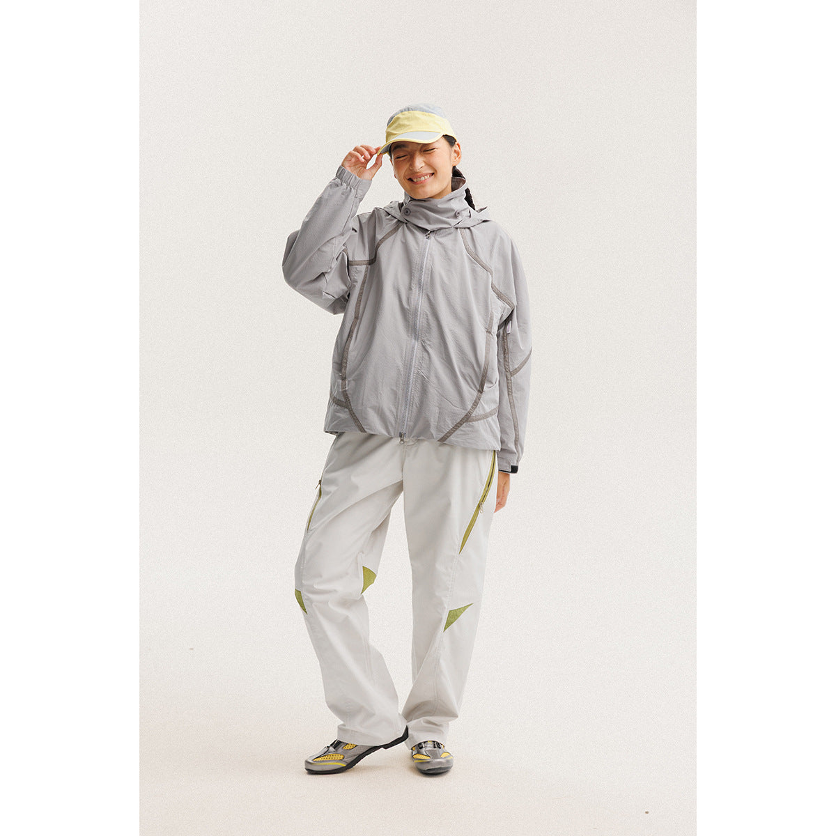 Model wearing a light gray hooded jacket and white pants with yellow accents, paired with a pale yellow cap.