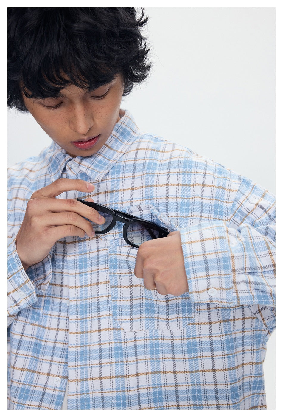 Close-up of a model holding sunglasses over a pale blue and tan plaid shirt with frayed chest-pocket edges.