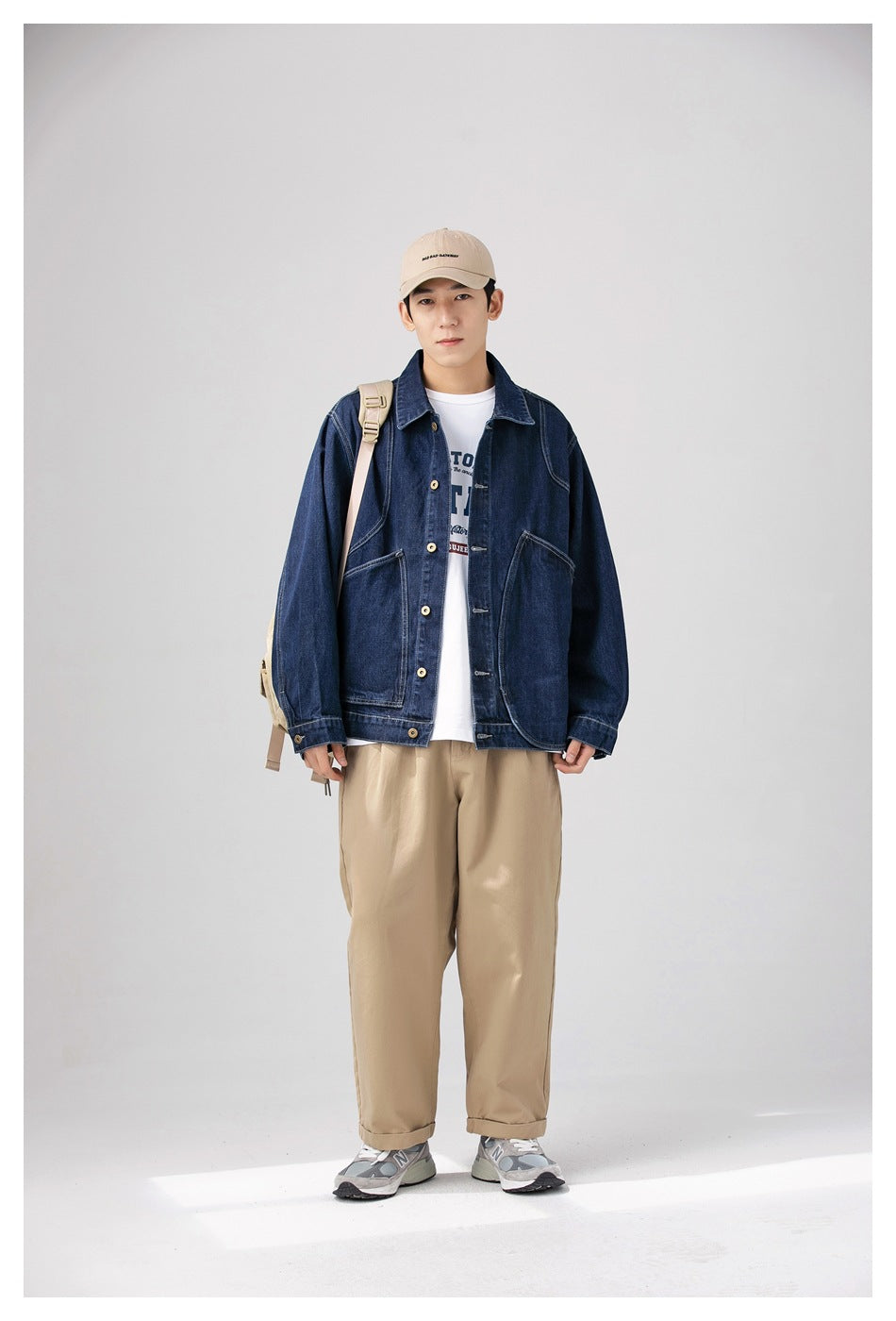 Model facing front in a dark denim jacket over a white graphic tee with khaki pants, beige cap and backpack