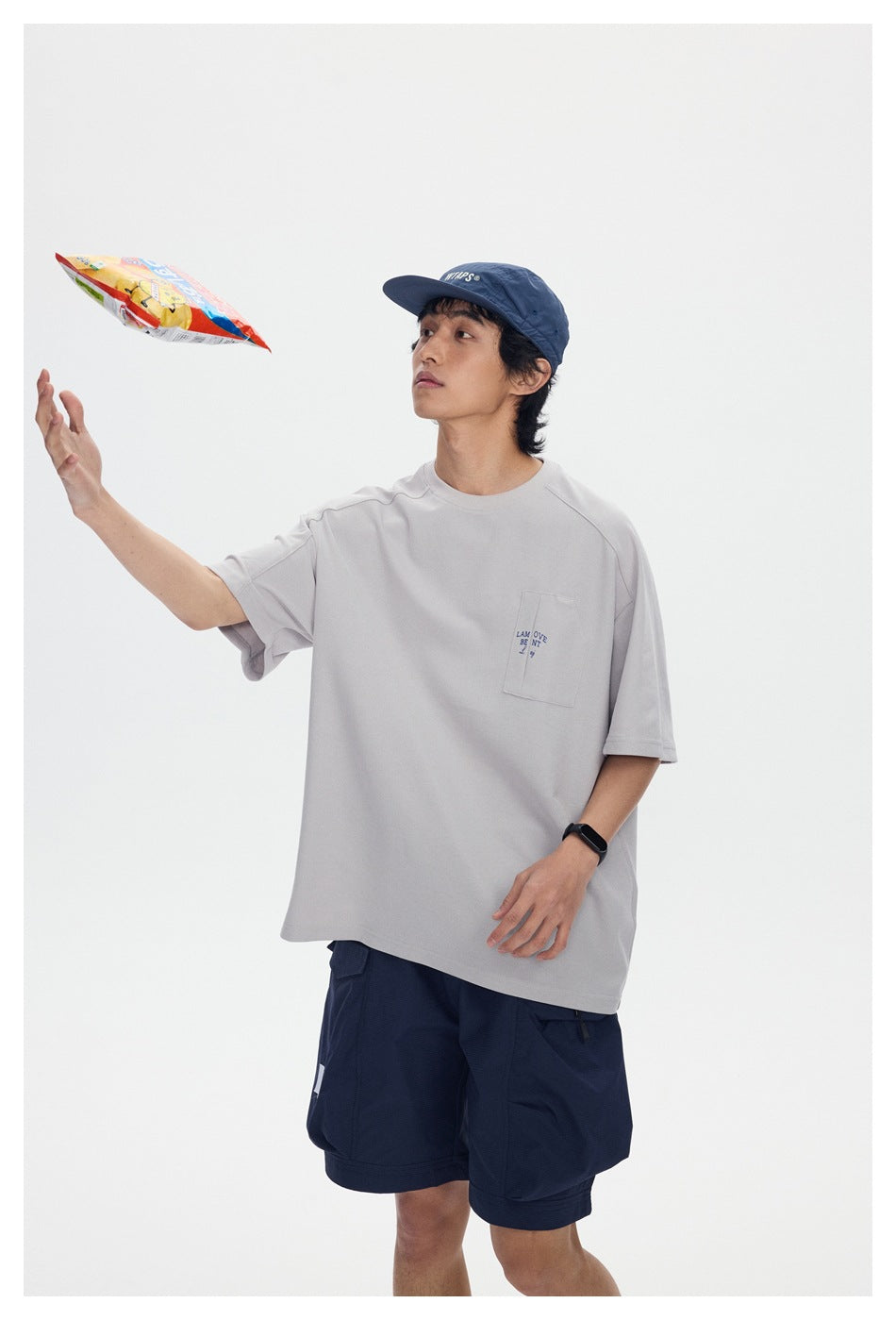 Model in a light gray pocket T-shirt, navy shorts, and cap tossing a snack bag.
