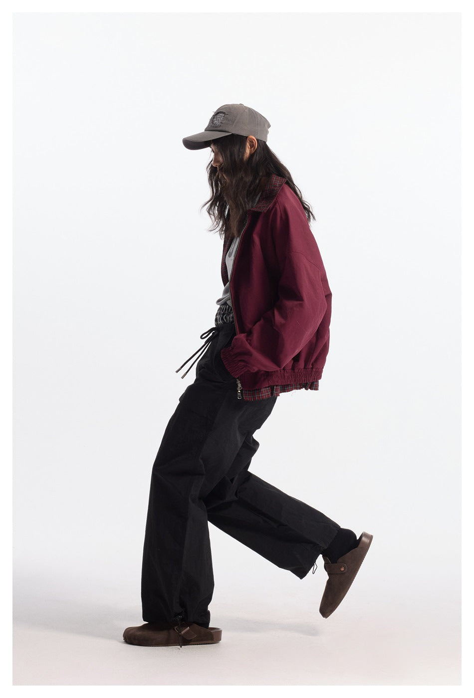 Side view of a model in a burgundy jacket with plaid trim, black trousers, and a gray cap.
