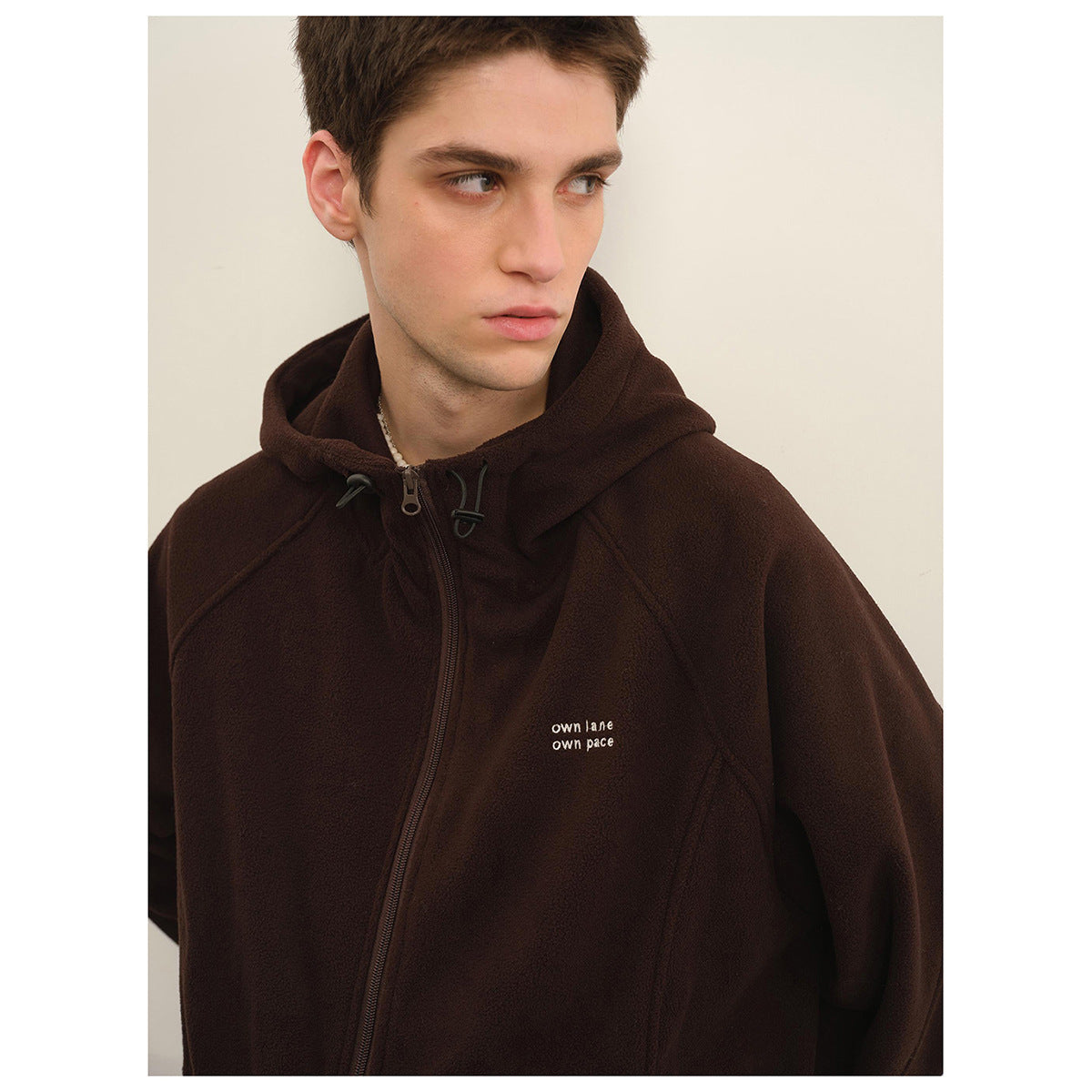 Close-up of a model wearing a dark brown zip-up hoodie with a drawstring hood and small white chest lettering.