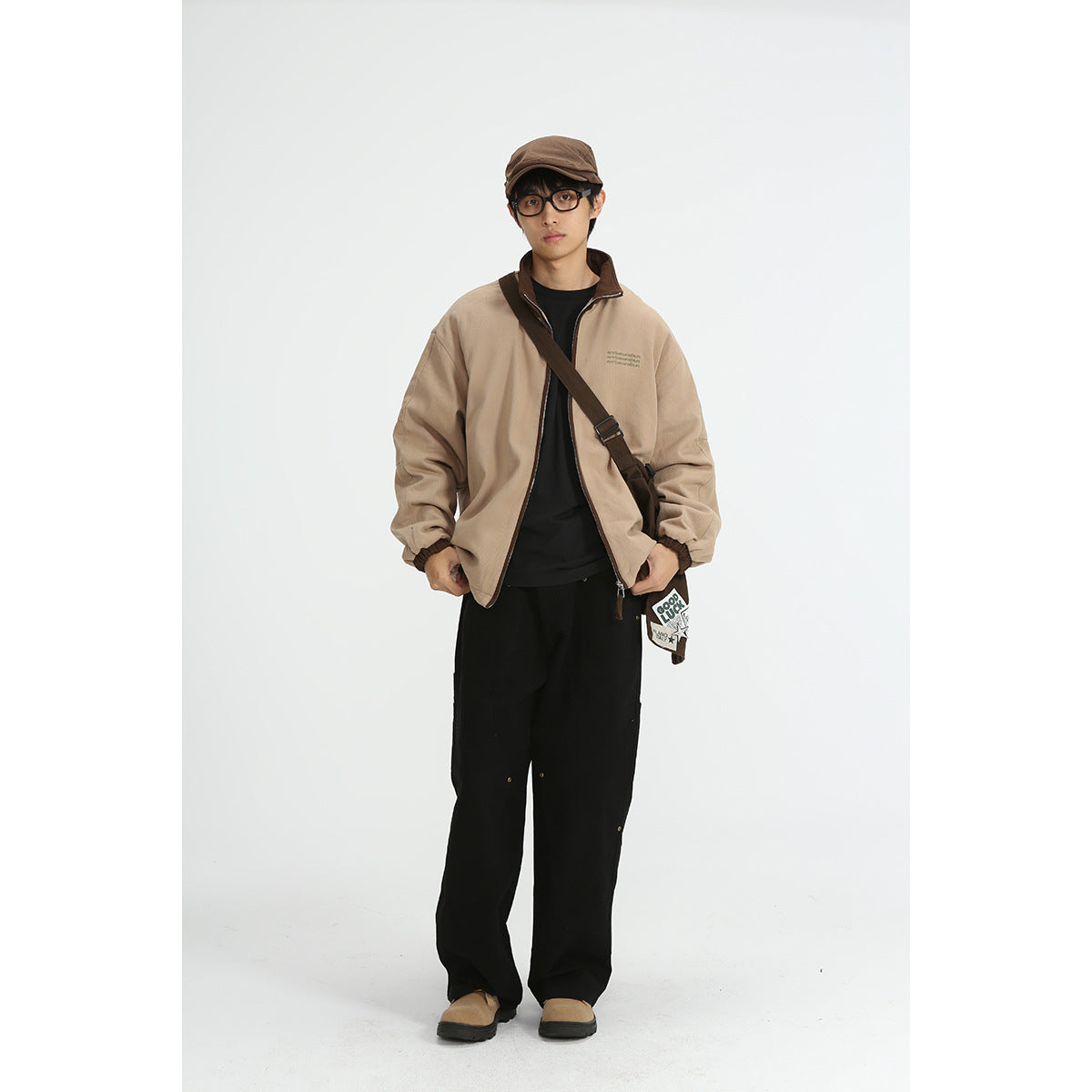 Model wearing a loose tan zip jacket with a dark brown collar, black pants, and a brown cap.