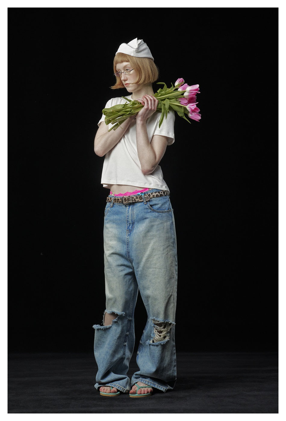 Model wears a white T-shirt and distressed wide-leg jeans, holding pink tulips across her shoulders against black.
