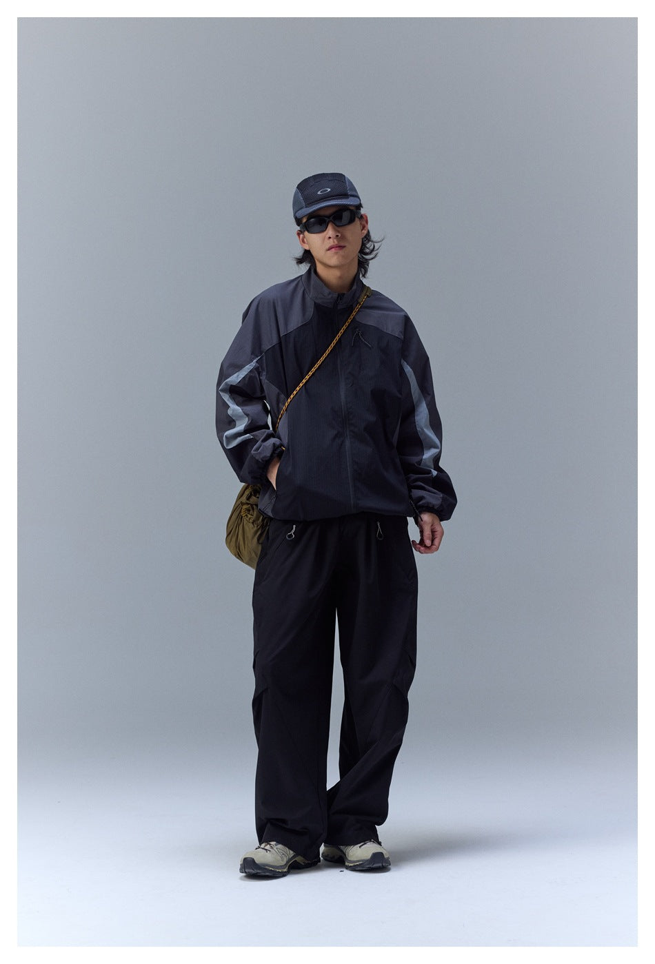 Full-length model wearing a black and gray outdoor jacket, loose black pants, cap, sunglasses, and trail shoes.
