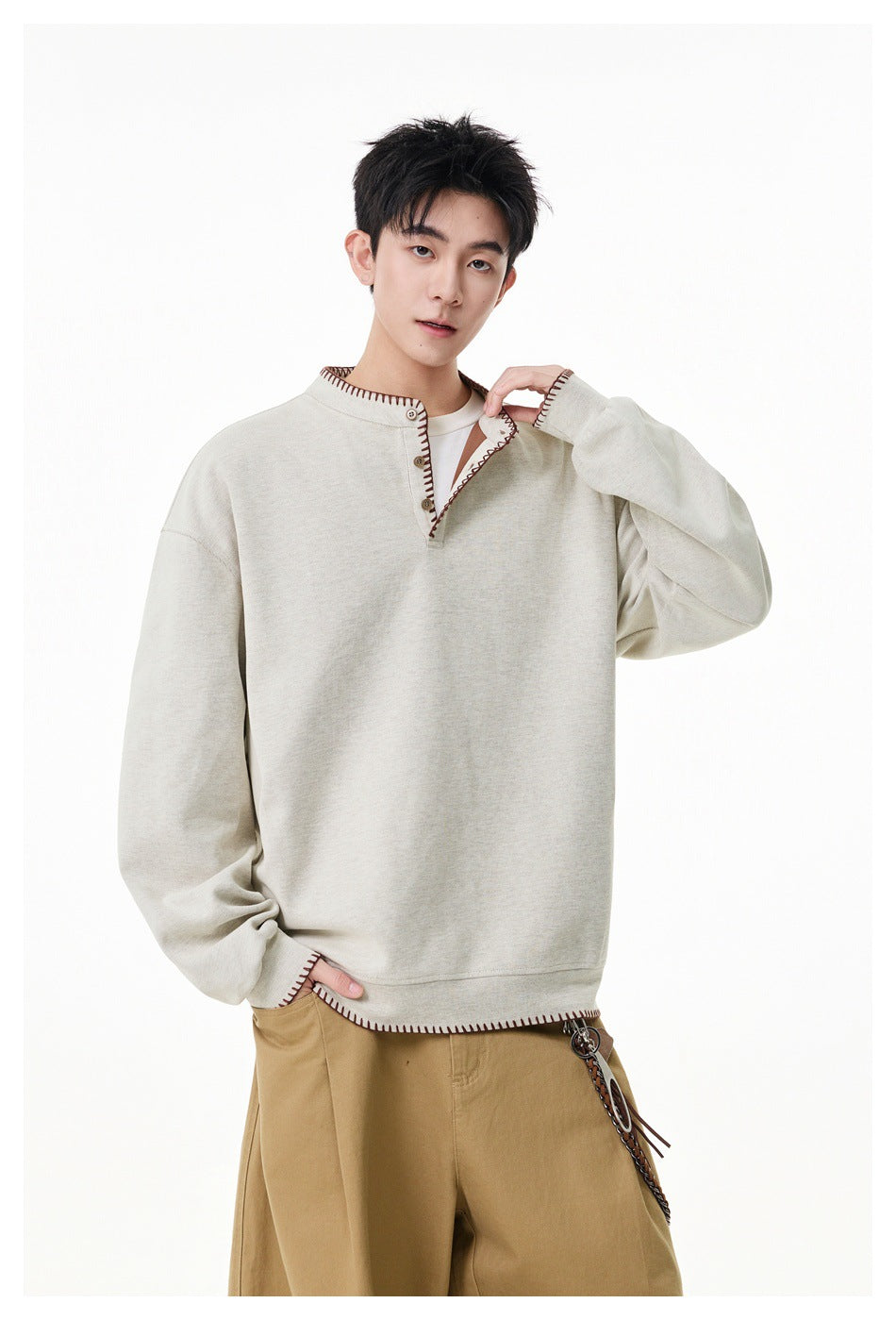 Model wearing a loose light beige long-sleeve pullover with a stand collar, button placket and dark contrast stitching.