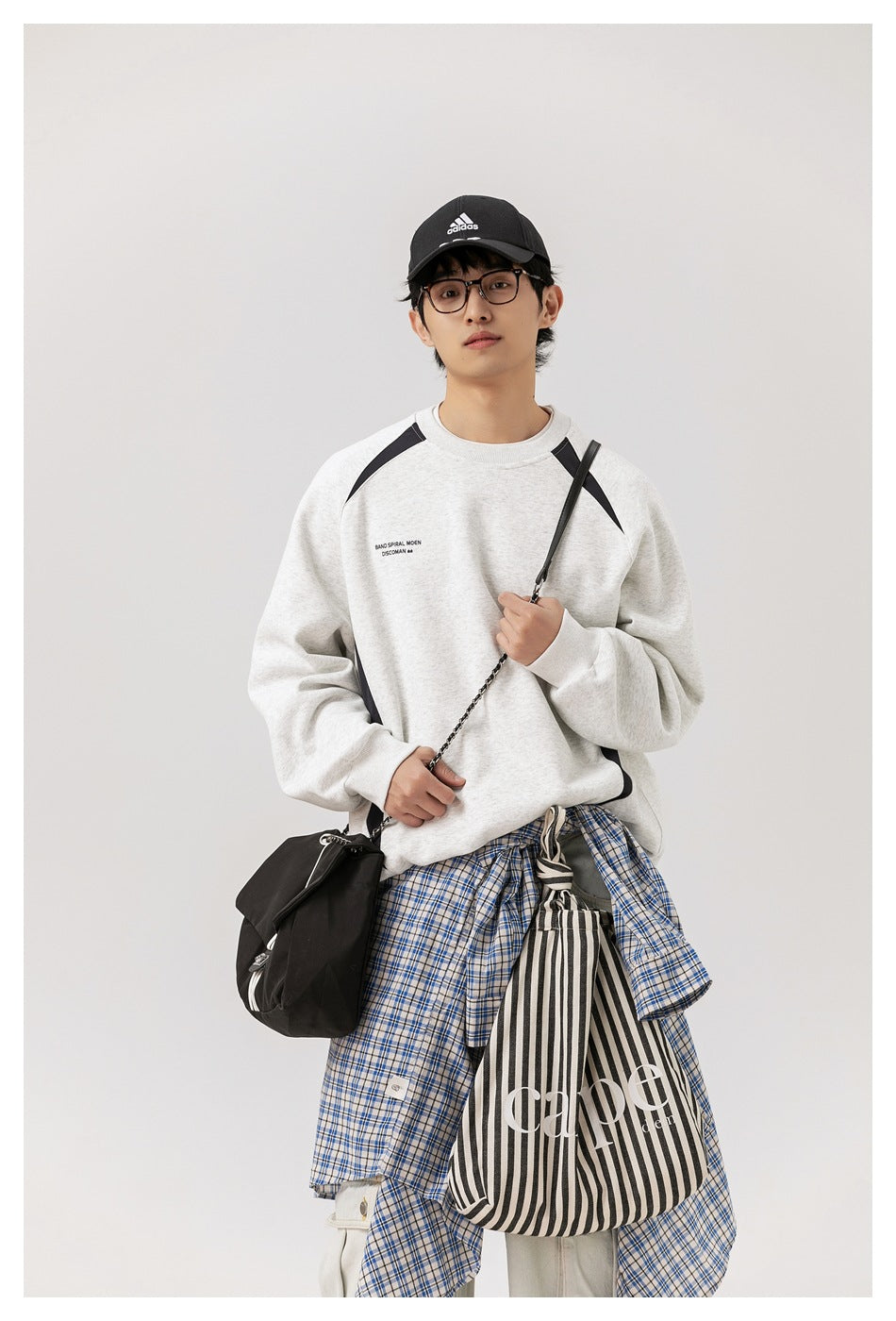 Model wearing a light gray sweatshirt with black curved panels, styled with layered bags and a blue plaid shirt.