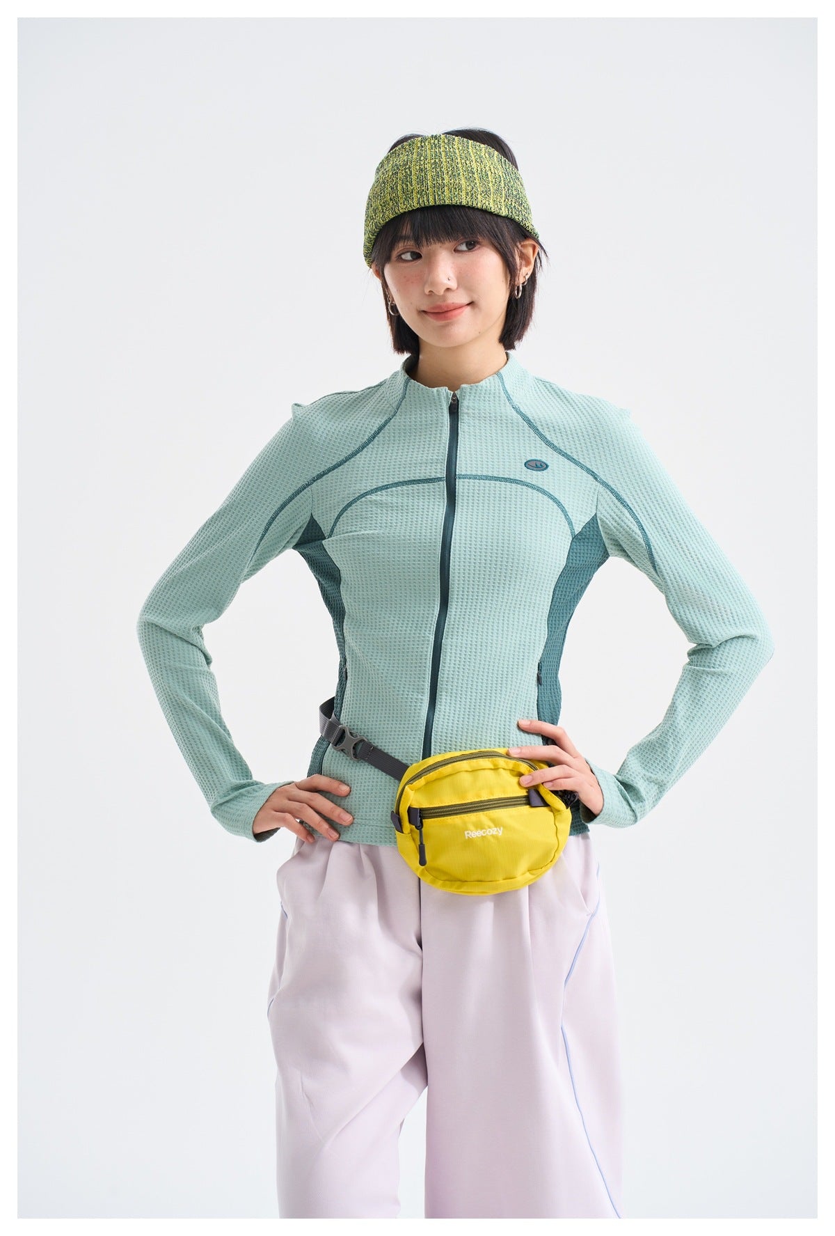 Model wearing a mint-green textured zip jacket with dark teal panels and a yellow waist bag.