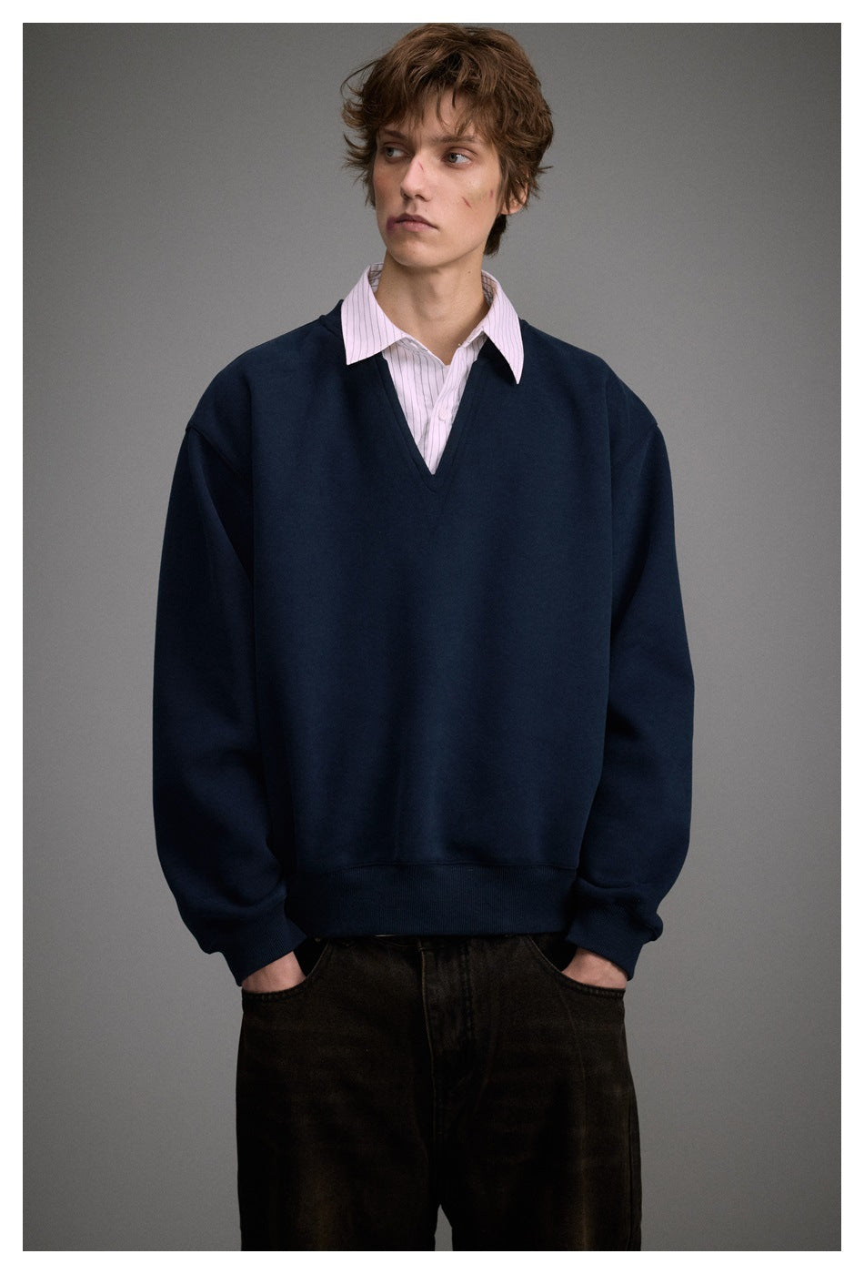 Model wearing navy V-neck sweatshirt over a pink striped collared shirt with dark pants, hands in pockets