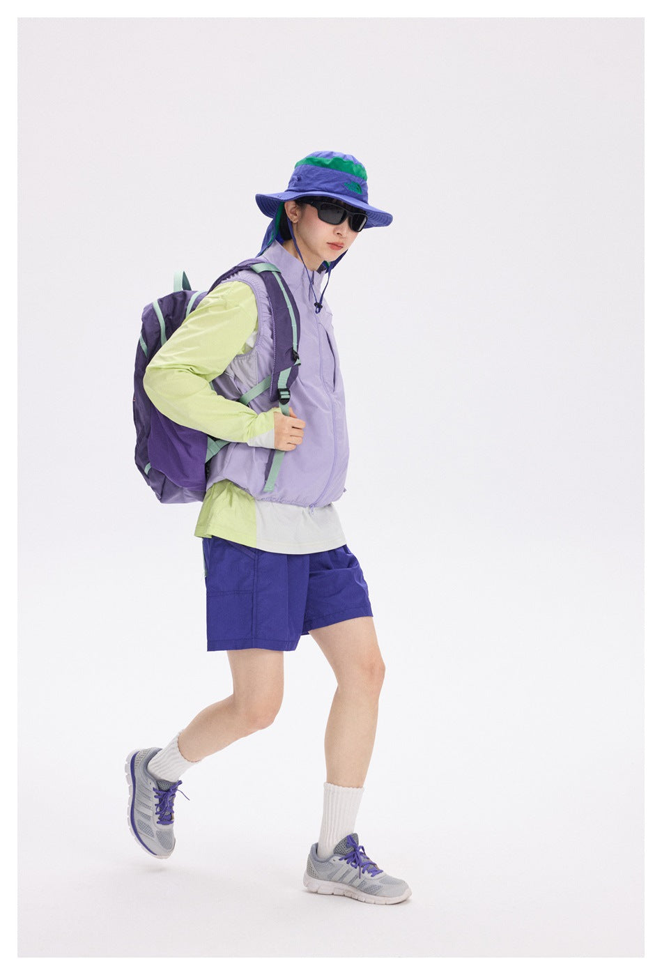 Model wearing a lavender vest, lime sleeves, purple shorts and hat, with a purple backpack and gray running shoes.