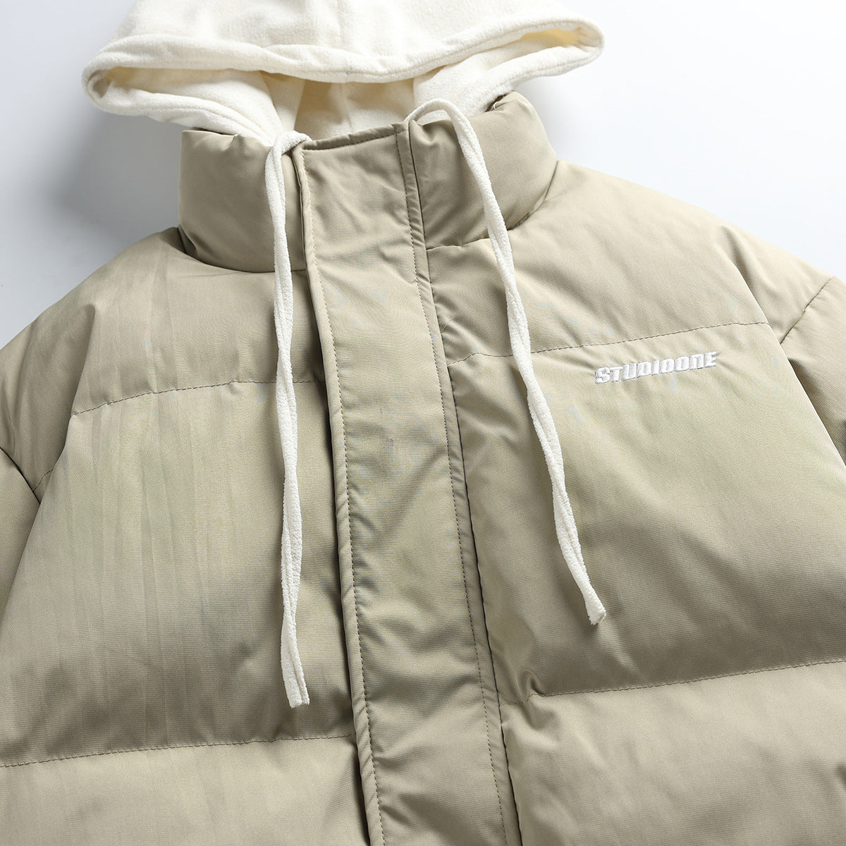 Close-up of a taupe puffer jacket front with a cream hood, drawstrings, high collar and white chest embroidery.