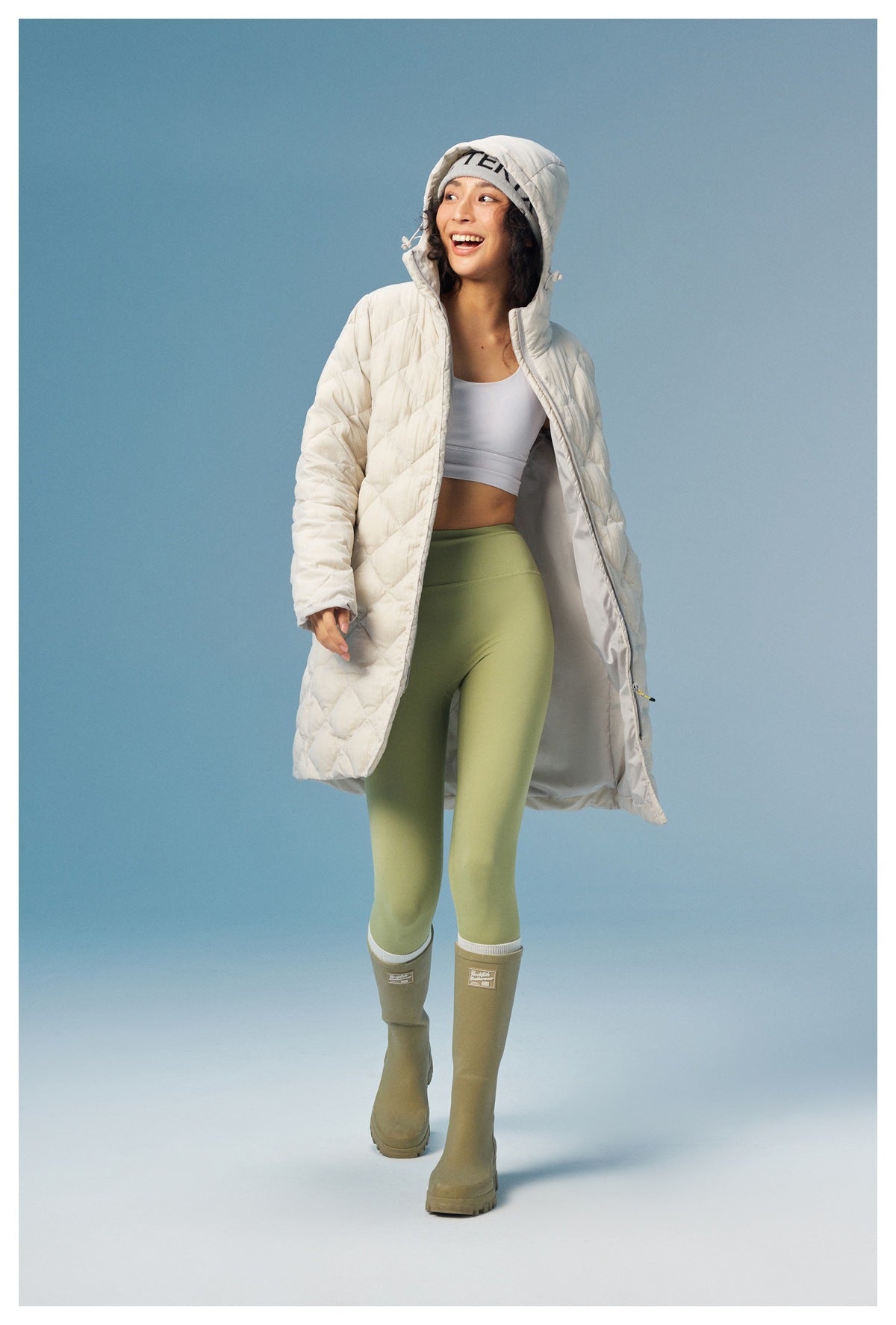 Model wears a cream quilted hooded coat open over a white top and green leggings, showing its light lining.