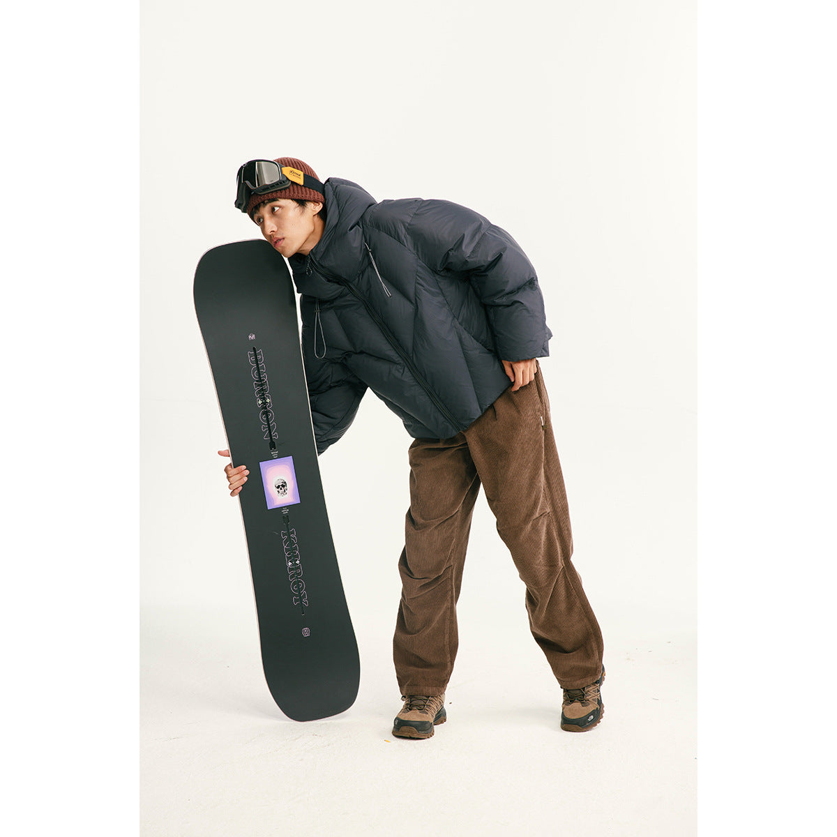 Model wearing a dark puffer jacket and brown pants beside a snowboard.