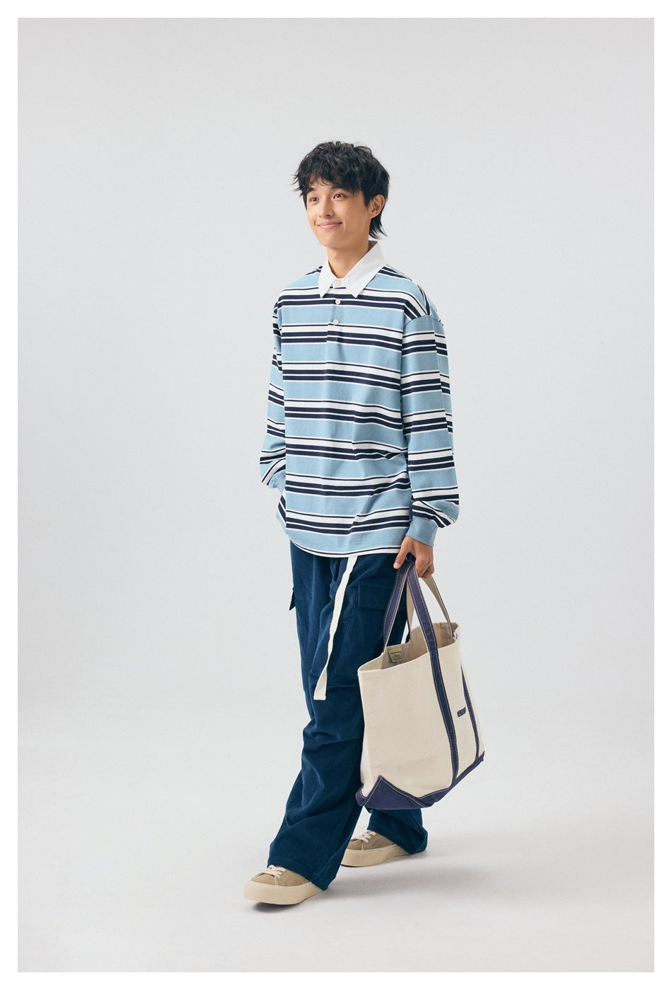 Full-length model wearing a light blue, navy and white striped rugby shirt and navy trousers, carrying a cream tote bag.